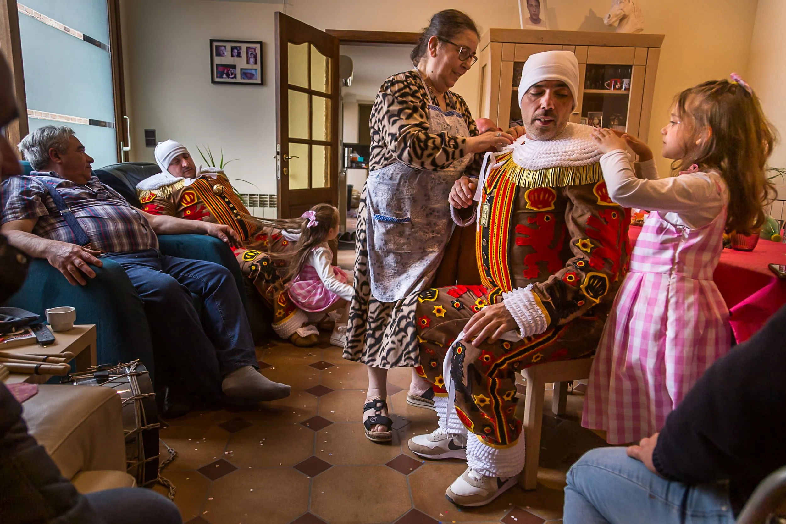 Une famille assise dans un salon, regardant une personne déguisée en roi avec un turban blanc, un costume coloré et des chaussures modernes, probablement lors d'une fête ou d'une célébration.