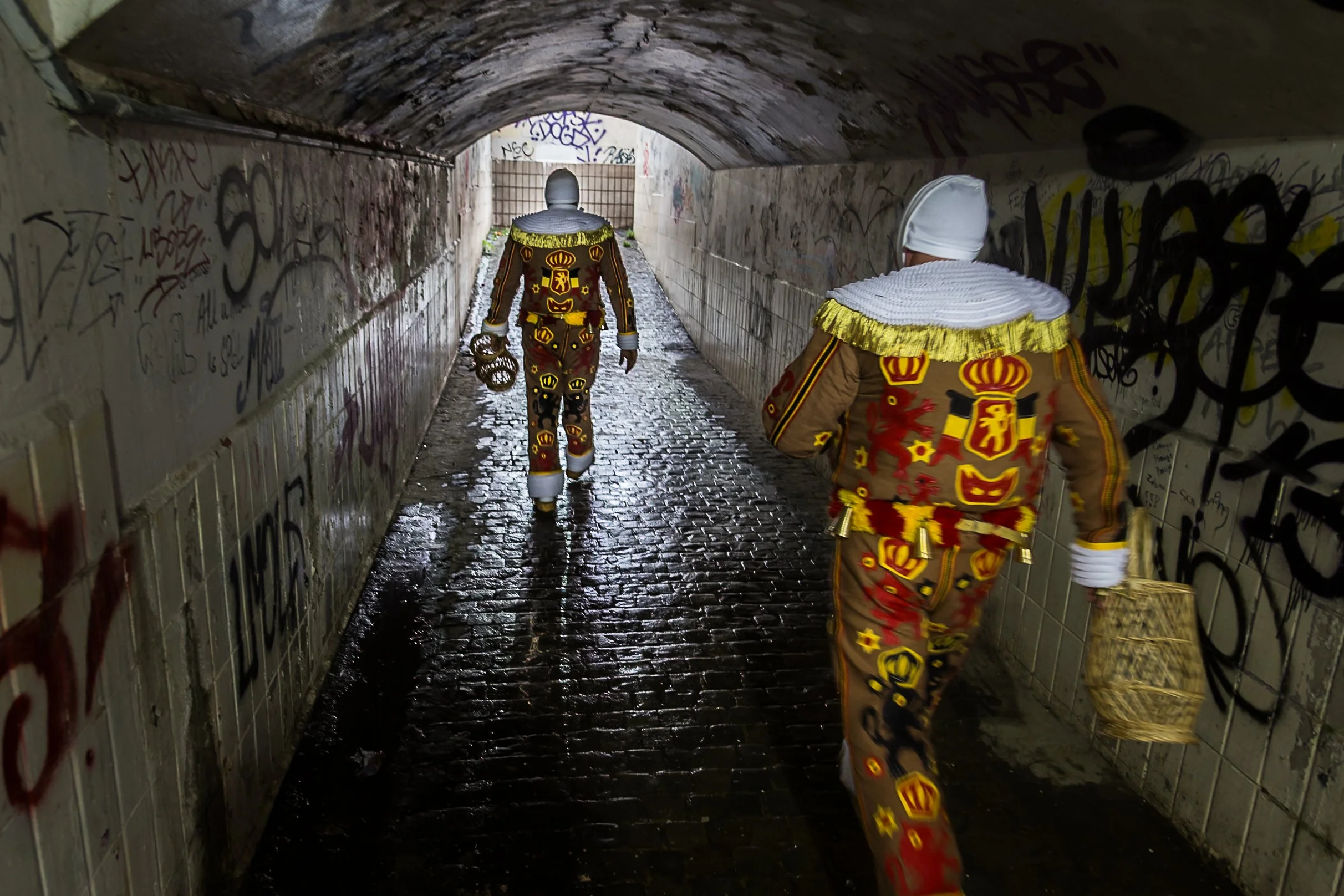Deux personnes vêtues de costumes traditionnels colorés marchent dans un tunnel graffité, au sol pavé humide.