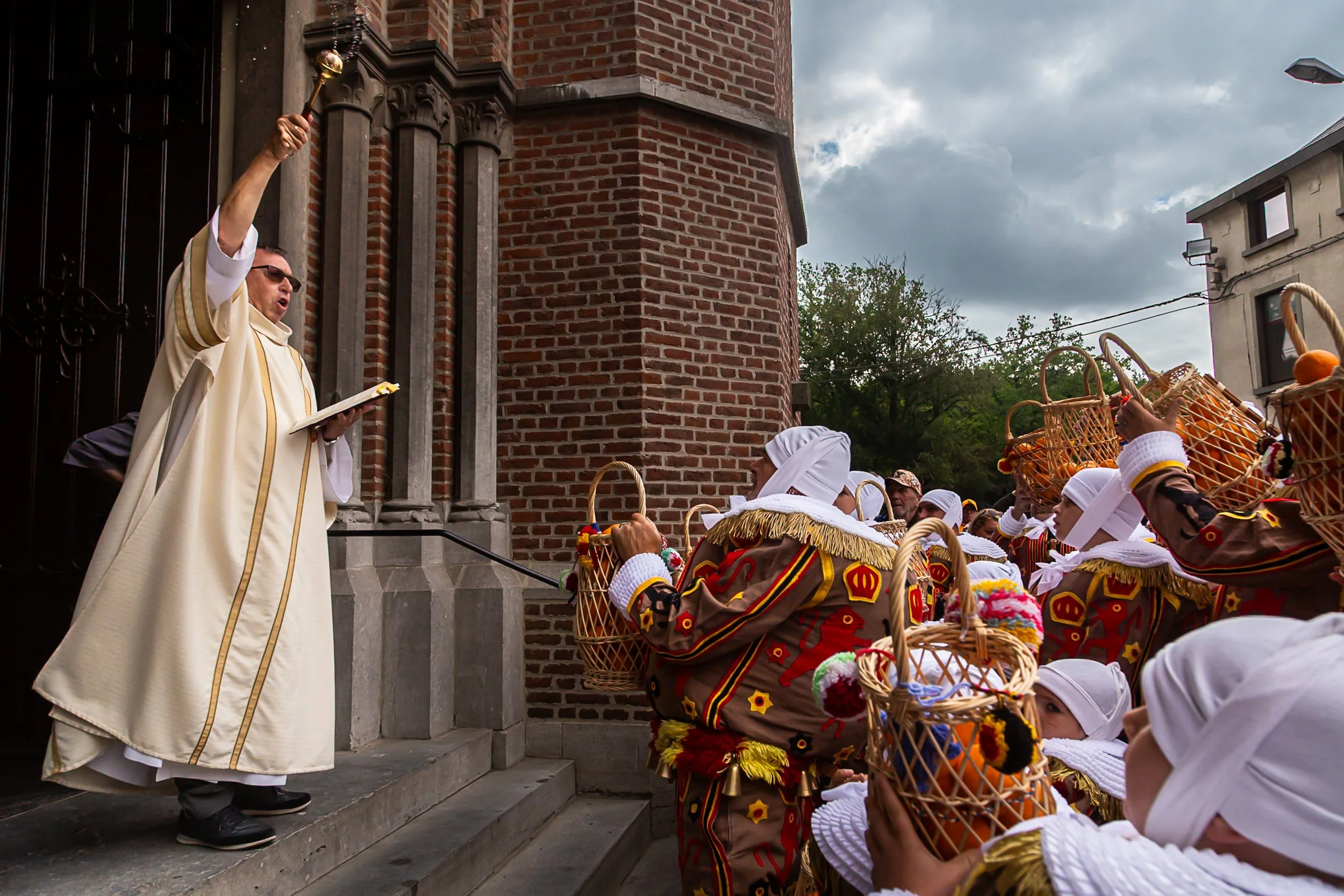 Un prêtre en robe blanche qui bénit une foule de personnes, notamment des enfants portant des costumes traditionnels colorés et des bonnets blancs, devant une église en pierre rouge, lors d'une cérémonie religieuse en plein air sous un ciel nuageux.