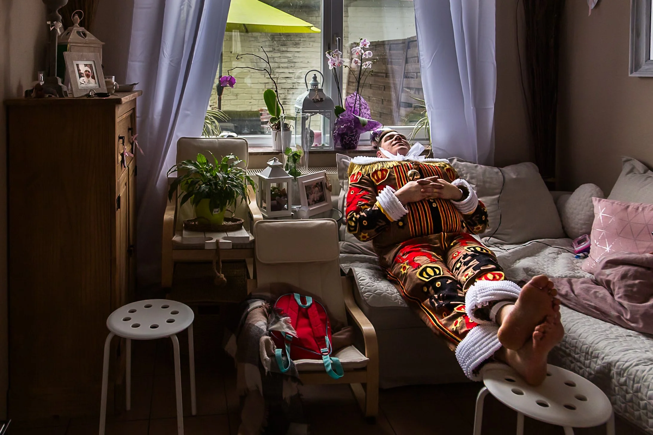 Une personne allongée sur un lit dans un salon, entourée de plusieurs plantes et décorations, avec une fenêtre en arrière-plan laissant entrer la lumière du jour.