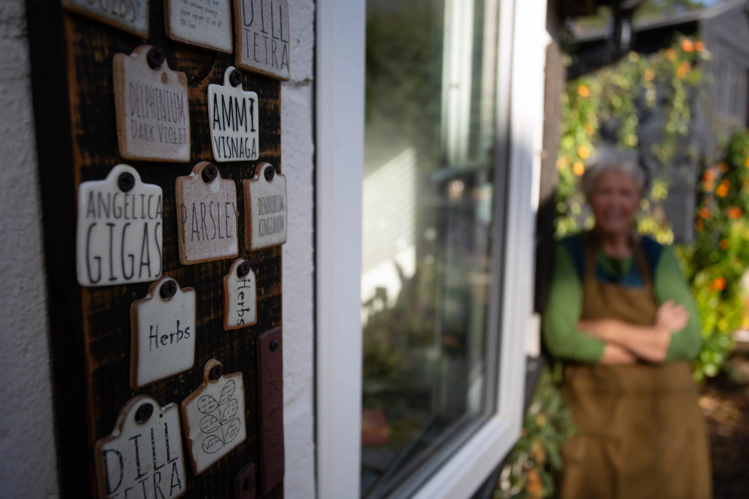 Handmade ceramic garden signs for herbs and flowers mounted on a wall, with ceramicist Sheena Bond in the background