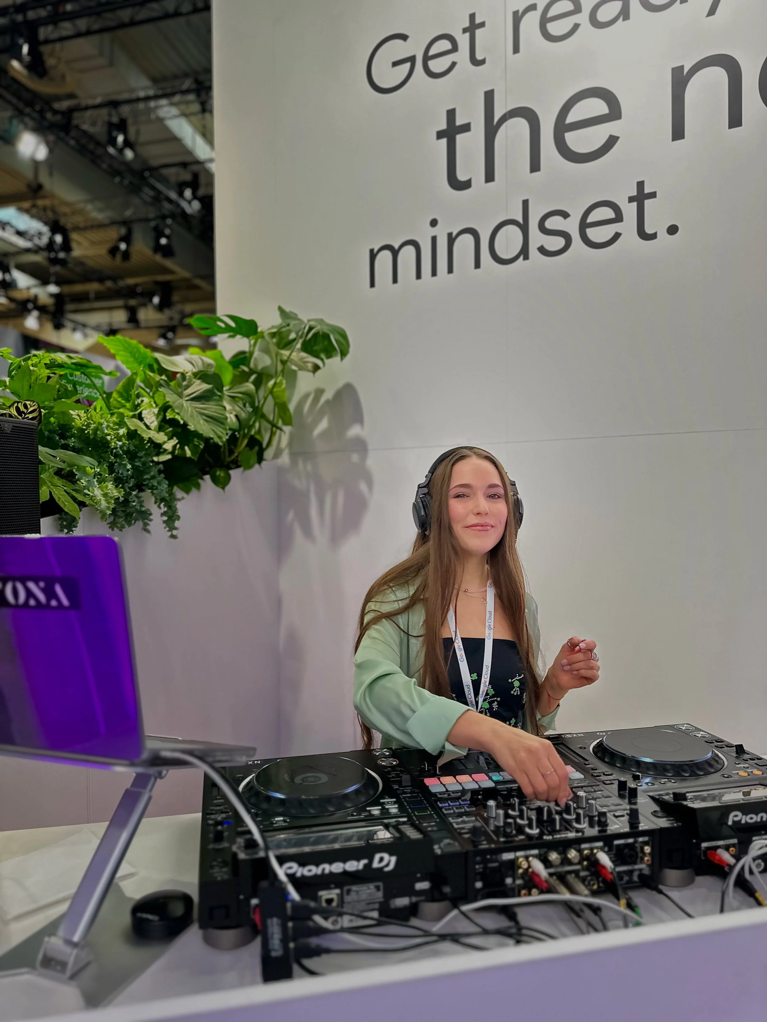 A young woman with long, light brown hair wearing headphones and a lanyard, smiling while DJing at a booth with Pioneer DJ equipment. There is a large white wall with the text 'Get ready, the n' and some green plants in the background.