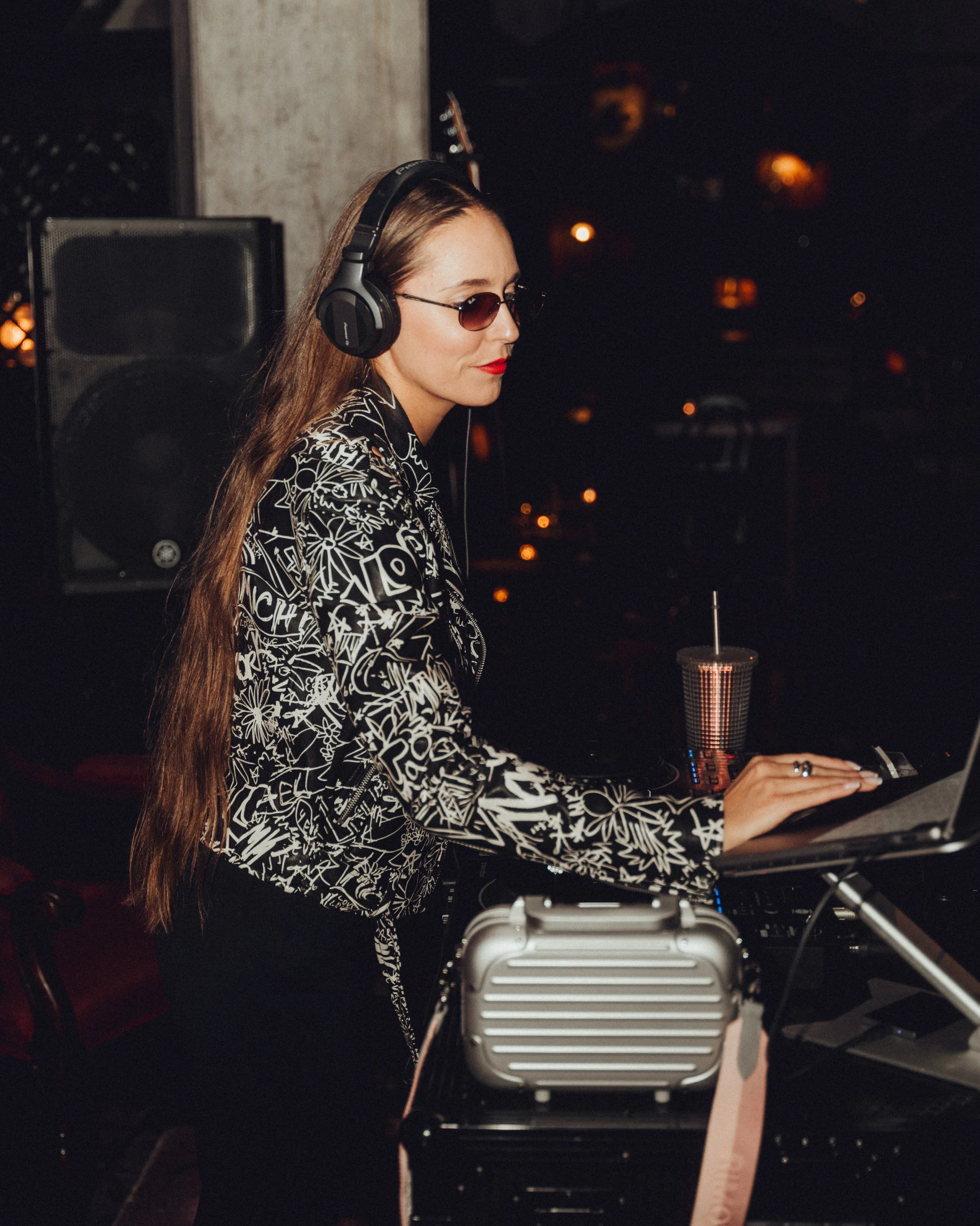 A woman with long hair wearing sunglasses, a patterned jacket, and red lipstick is DJing at a dark venue with warm lighting, surrounded by audio equipment and a cup with a straw.