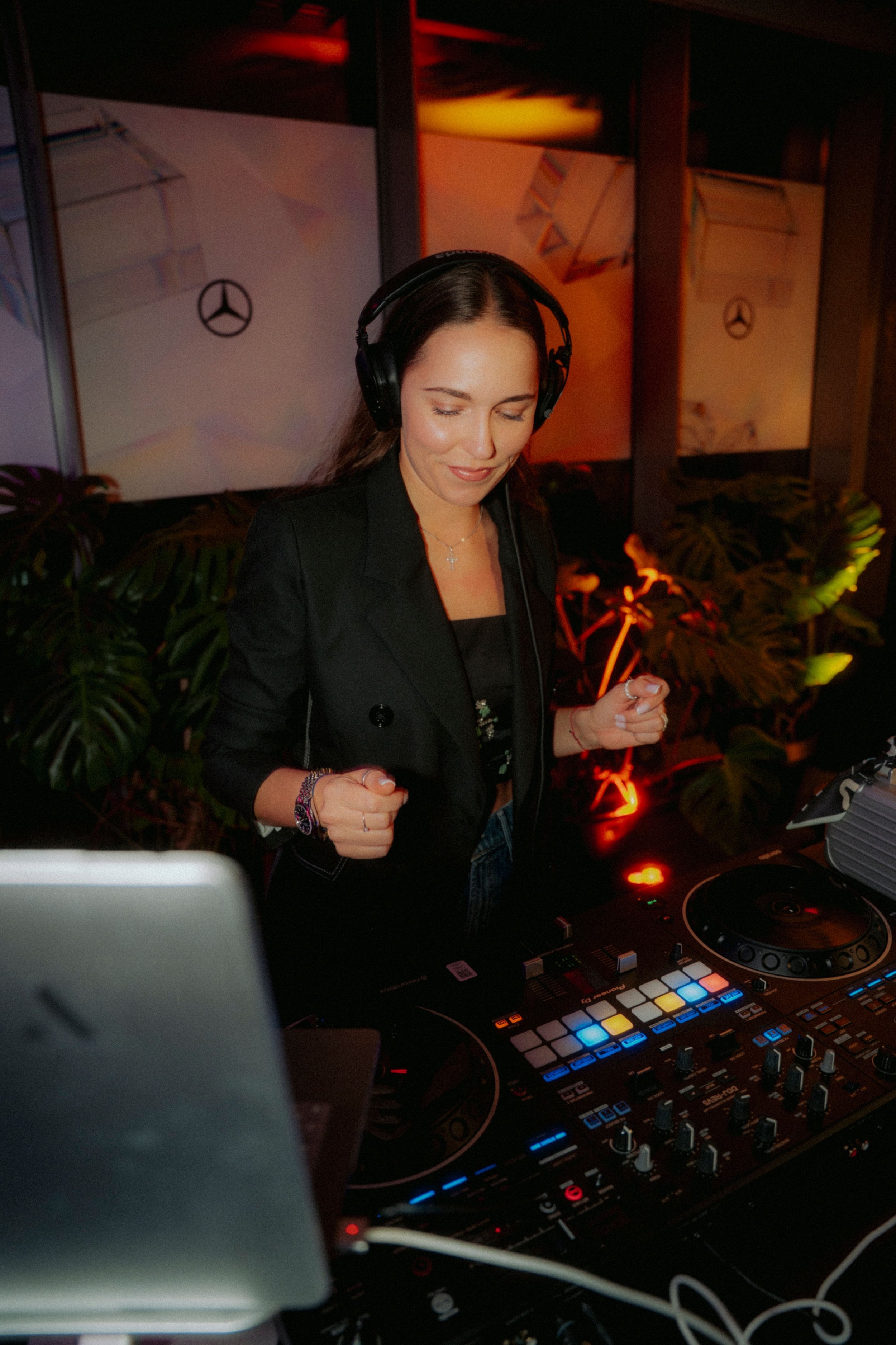 A female DJ with long brown hair wearing headphones, a black blazer, and a cross necklace, is smiling and dancing while operating DJ equipment at a lively event with colored ambient lighting and green plants in the background.