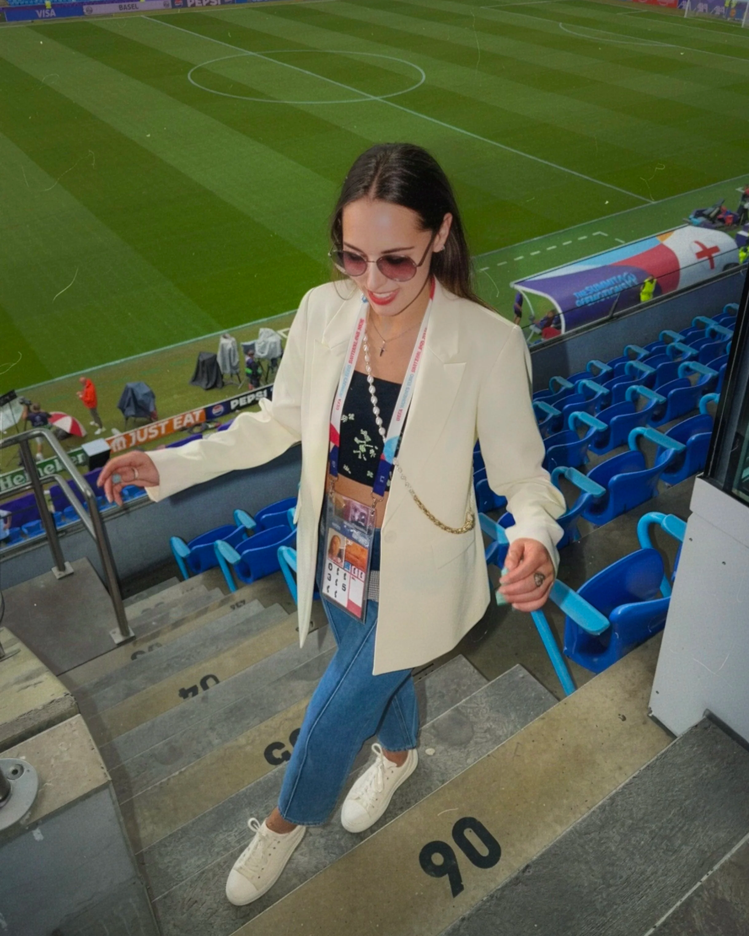 A woman taking a selfie in front of a mirror at a soccer stadium. She is wearing sunglasses, a white blazer, a black top, blue jeans, and white sneakers. The stadium seating and a soccer field are visible in the background.