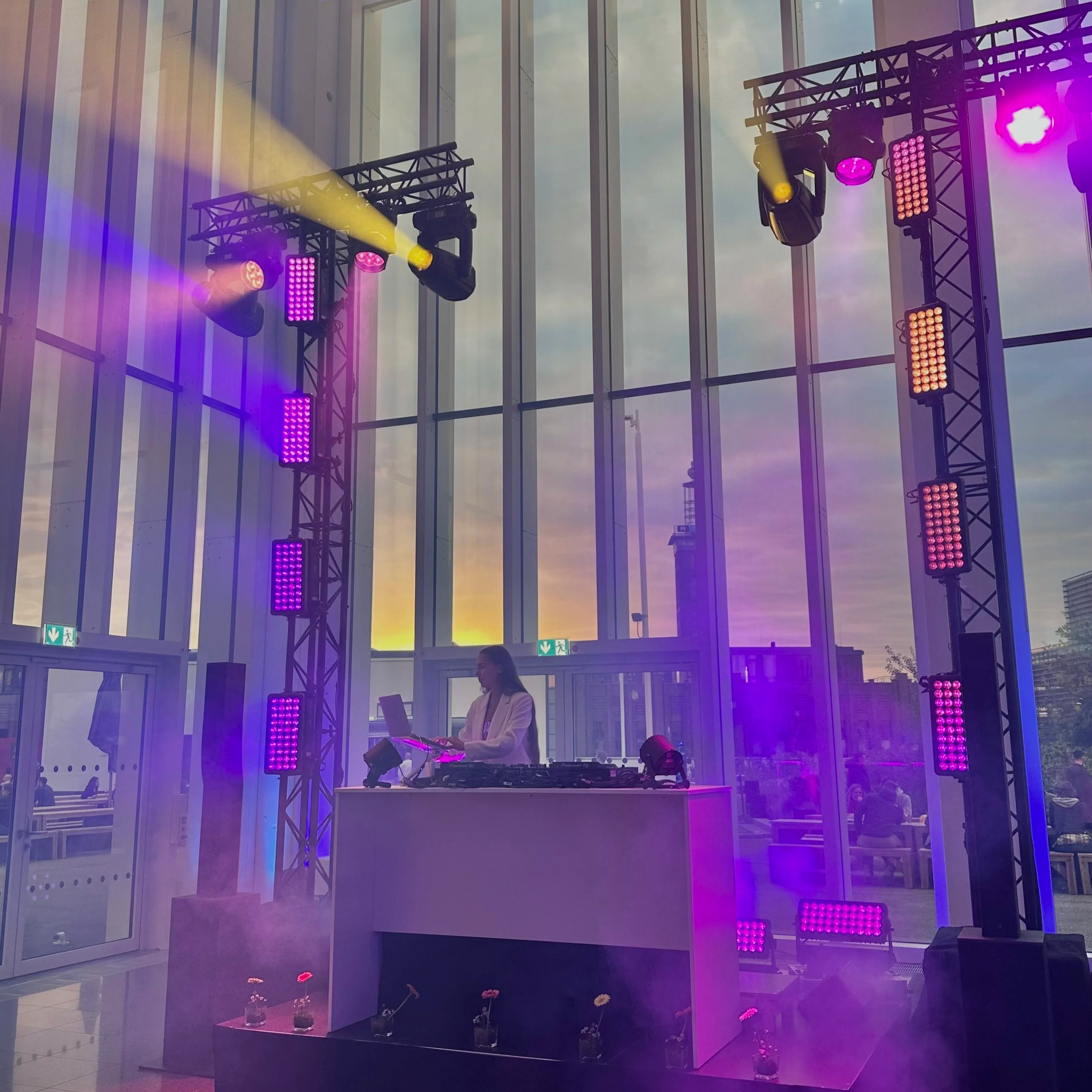 Indoor event space with a DJ booth, purple and yellow stage lighting, and flowers in small vases on the floor