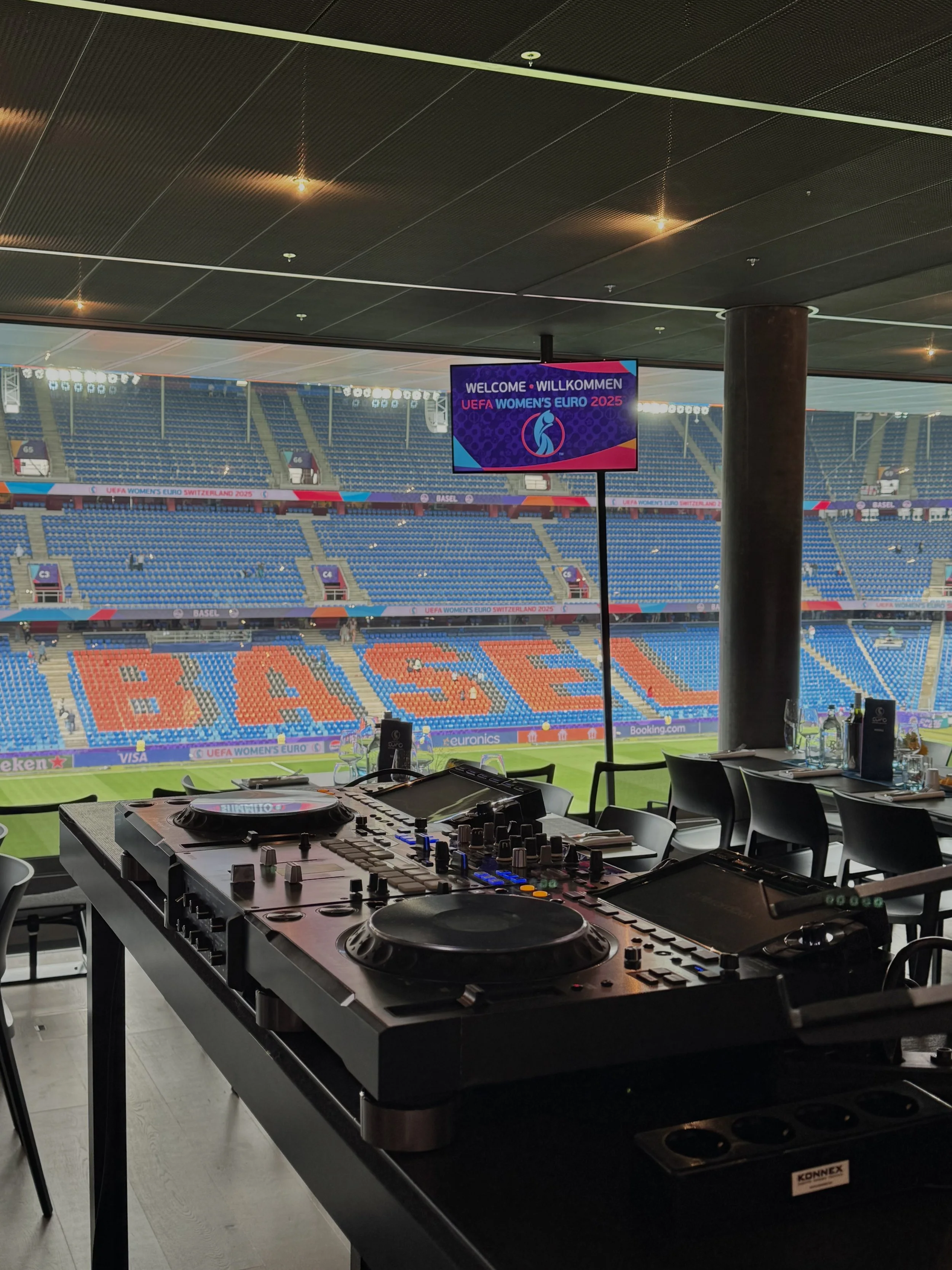 DJ equipment on a table overlooking a stadium with empty seats and a large screen displaying a message about the UEFA Women's Euro 2025 in Switzerland, with a seating area spelling out 'BASEL' in orange and blue seats.
