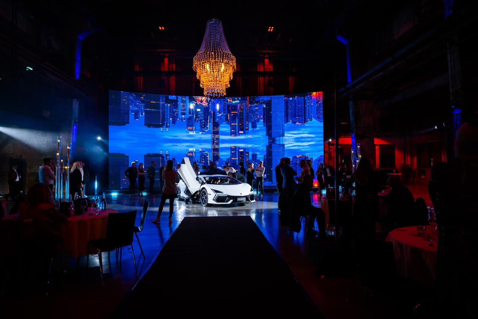 A white luxury sports car with doors open displayed on a stage at a technology or car event, with a large digital cityscape backdrop and people observing the presentation, illuminated by colorful lights and a chandelier.