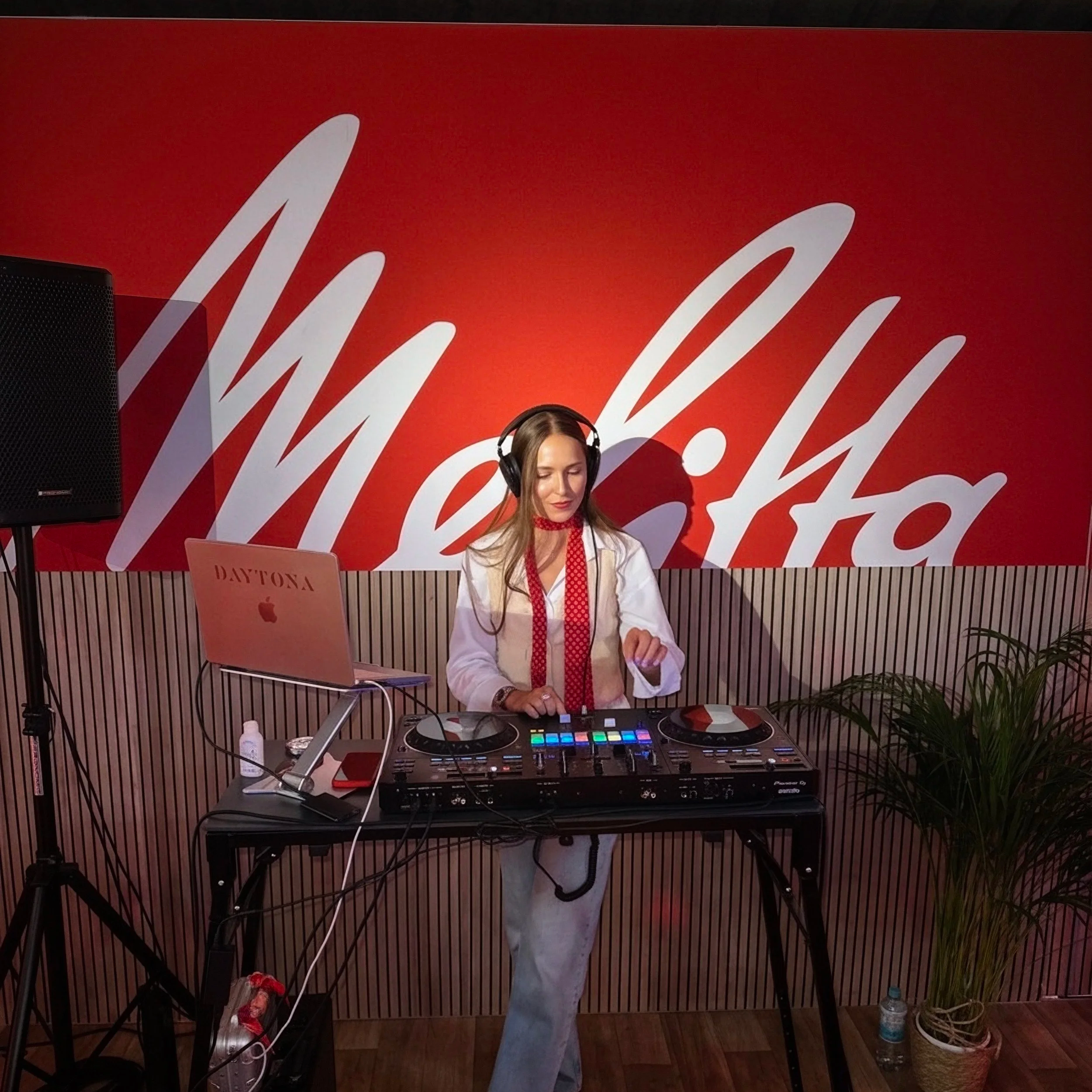 A woman with long hair wearing headphones, white jacket, and red tie, standing behind DJ equipment with colorful lights, in front of a red wall with white text, and a laptop with 'DAYTONA' branding.
