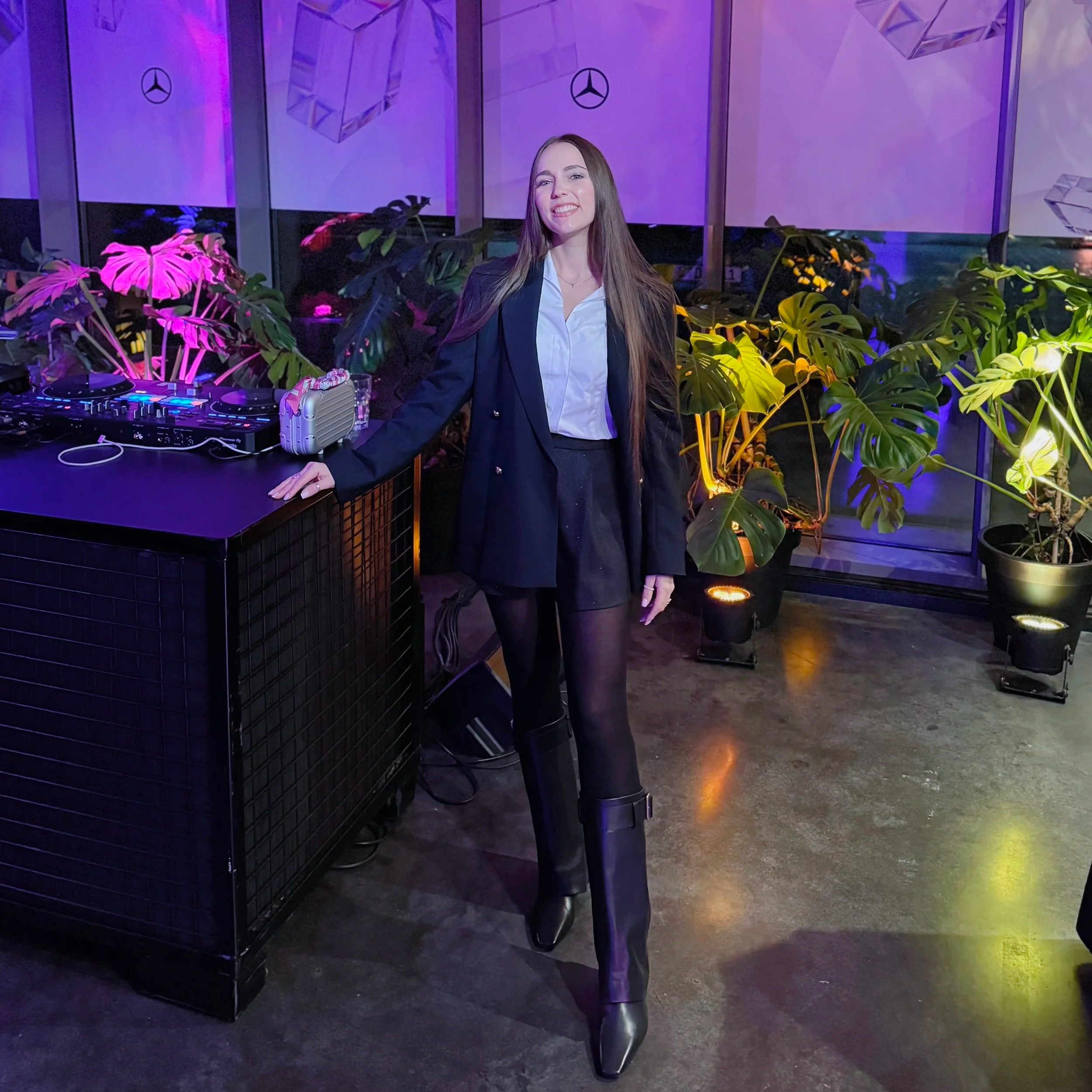 A woman with long brown hair wearing a black blazer and white shirt standing near a bar counter with DJ equipment in a nightclub setting. The background features purple and yellow lighting and large patterns on the walls with Mercedes-Benz logos. There are various plants around her, including Monstera and other large-leafed varieties.