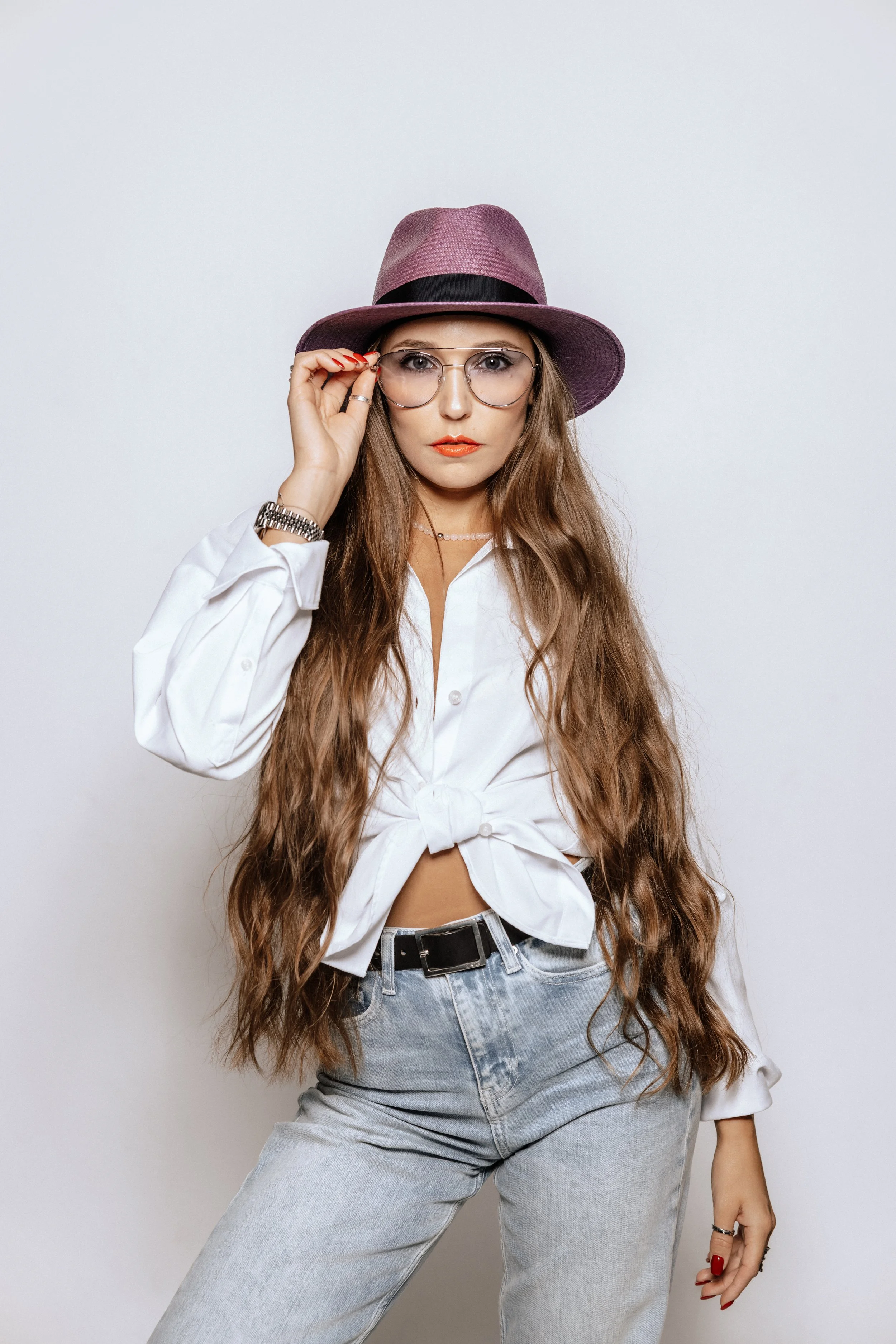 A young woman with long wavy hair wearing a purple wide-brimmed hat, clear glasses, a white tied-up shirt, light wash jeans, and a watch, standing against a plain gray background.