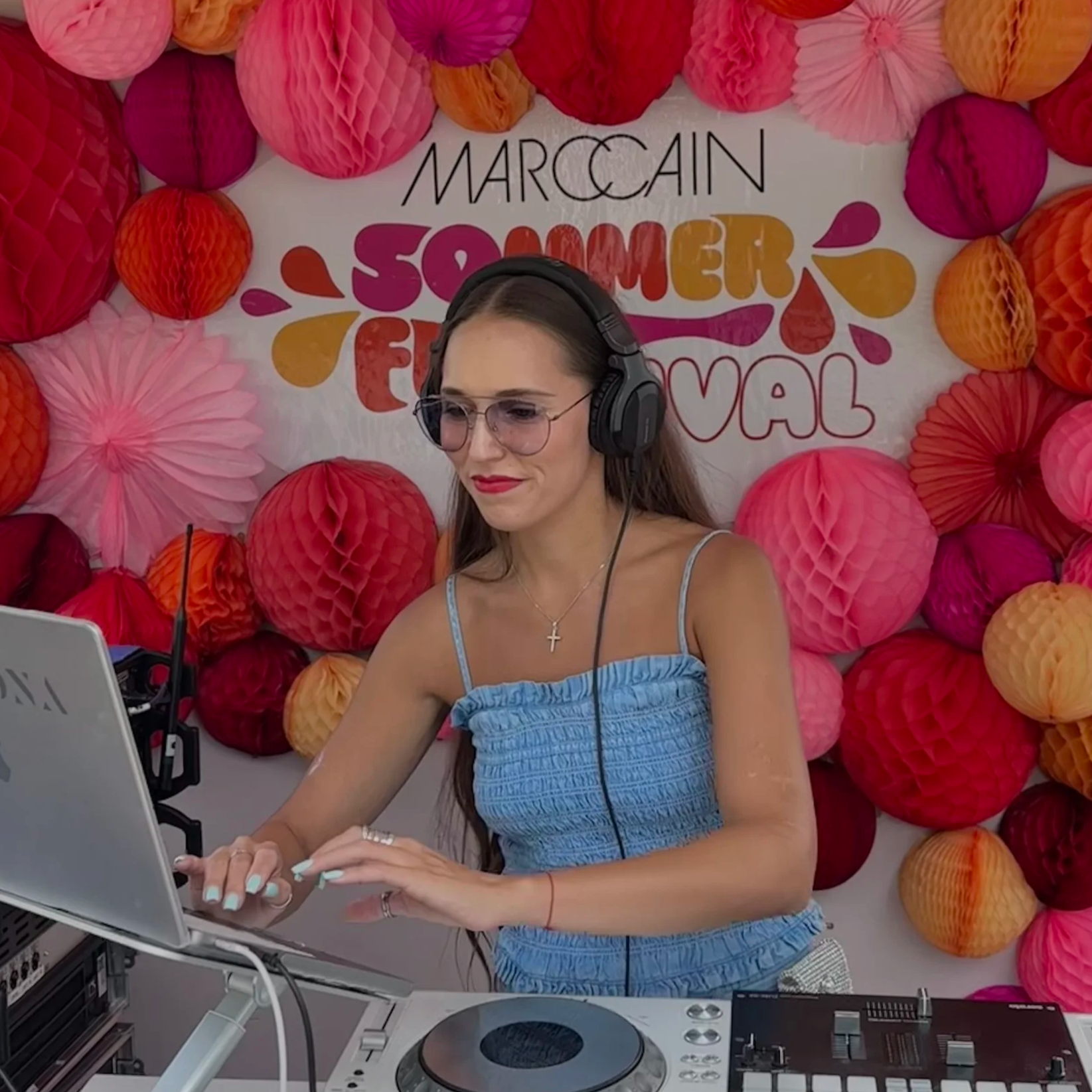 A woman with long brown hair, wearing glasses, a cross necklace, and a blue sleeveless top, working on a laptop at a DJ booth with colorful paper lantern decorations and a sign that reads 'Marccain Summer Festival' in the background.
