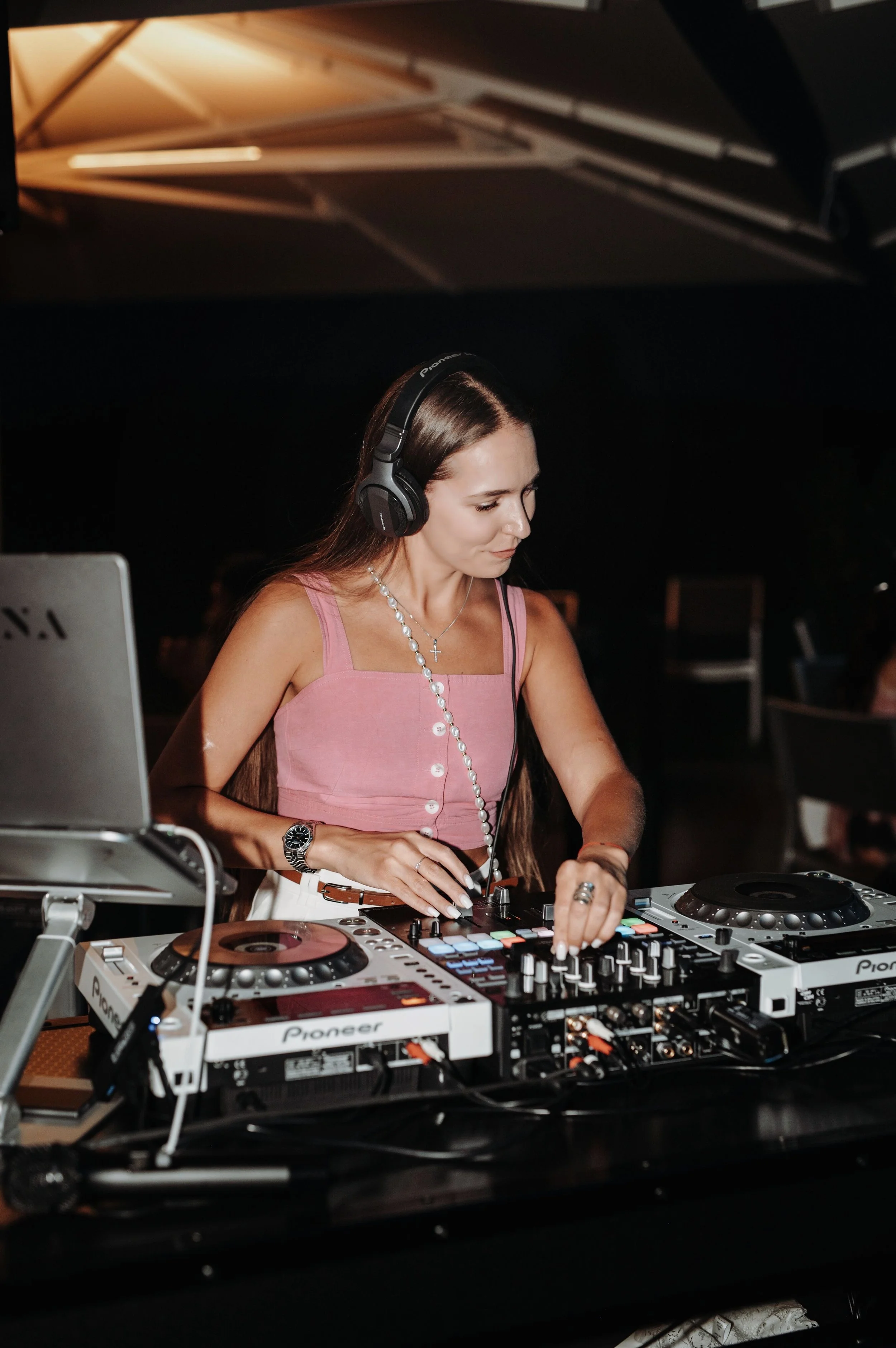 A female DJ wearing headphones and a pink sleeveless top, operating professional DJ equipment with colorful buttons and turntables in a dark setting.