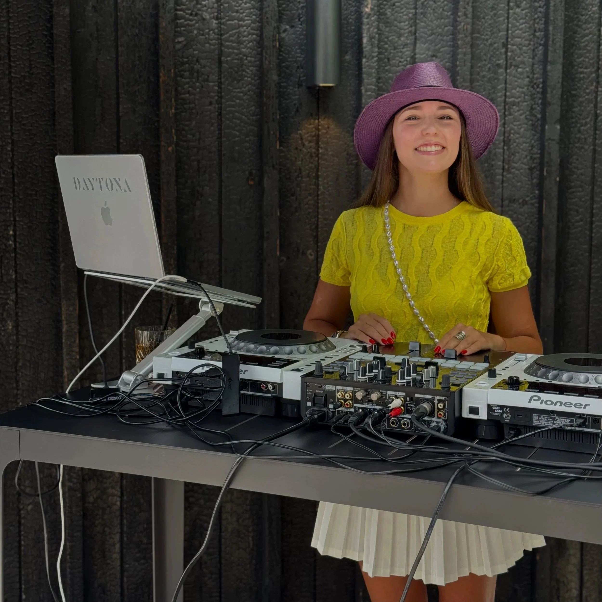 A woman with long brown hair wearing a purple wide-brimmed hat and yellow dress, standing at a DJ booth with music equipment, against a dark wood-paneled wall.