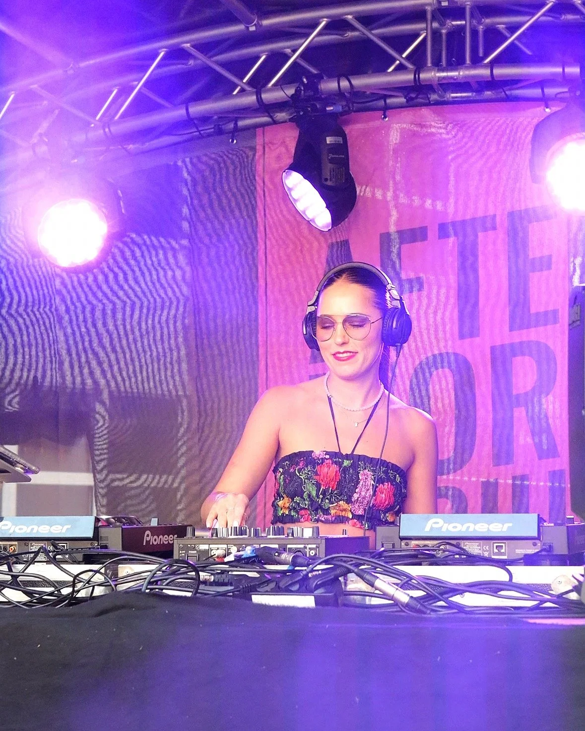 A female DJ with sunglasses and headphones performing at a club, with purple lighting and a large pink banner in the background.
