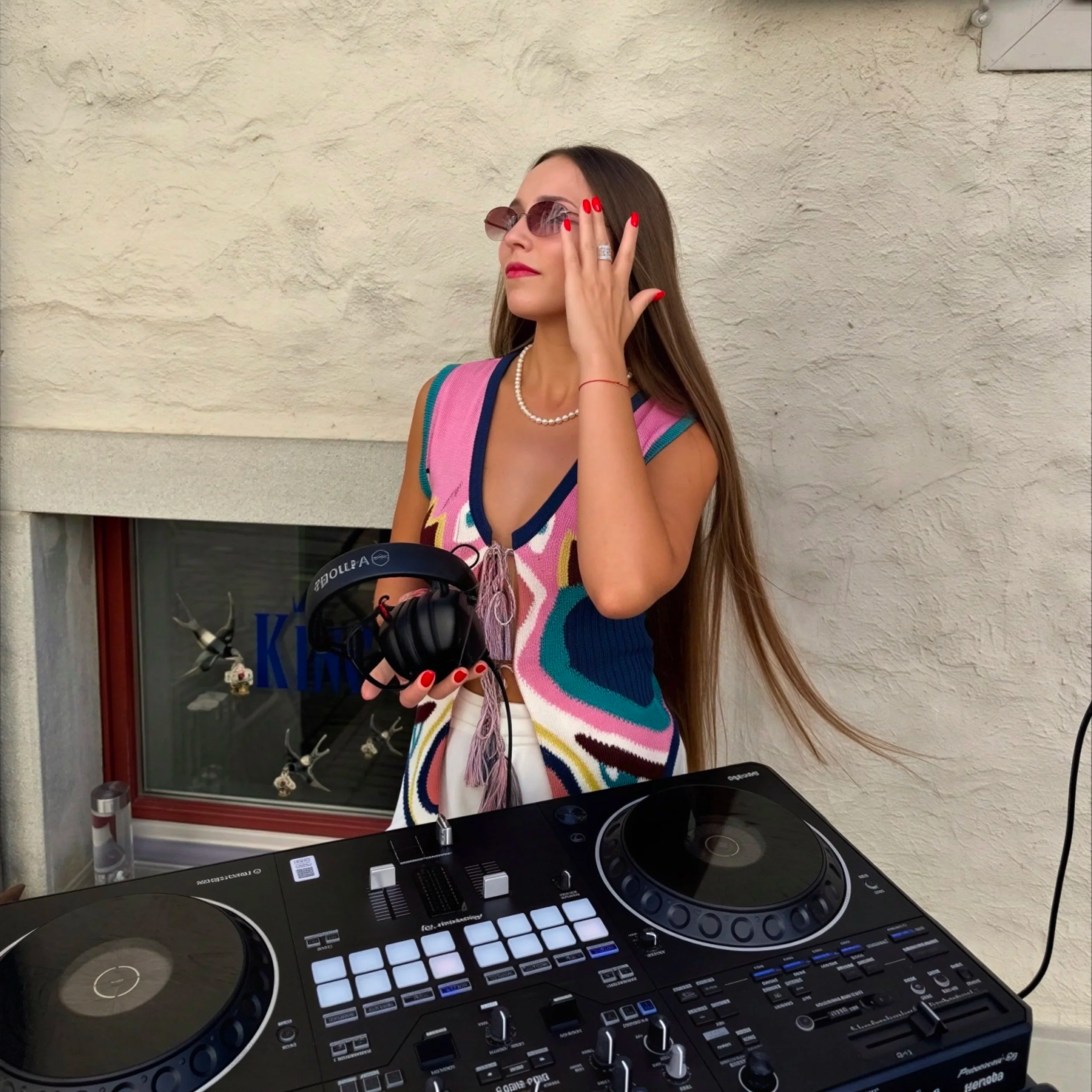 A woman with long brown hair wearing sunglasses, a pearl necklace, and a colorful sleeveless top stands outdoors against a beige textured wall. She has red nail polish and is touching her sunglasses with her right hand.