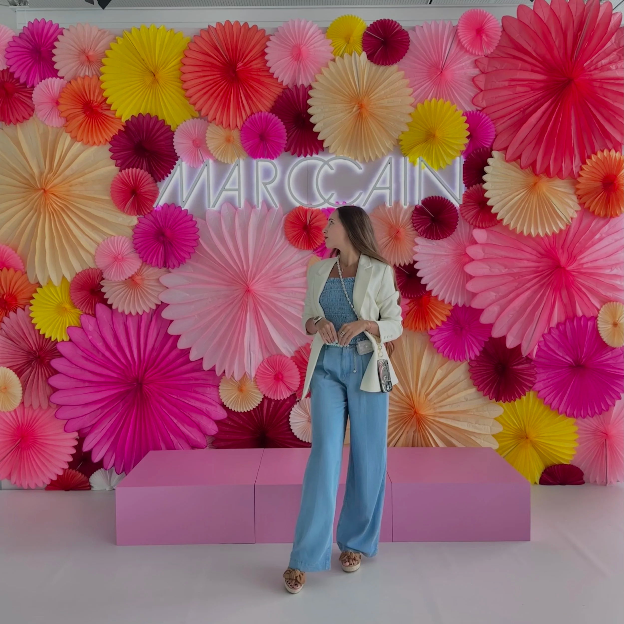 Person standing in front of a colorful backdrop decorated with large paper fans in pink, yellow, red, and cream colors. The person is wearing a white blazer, light blue jeans, and sandals, and appears to be looking to the side.