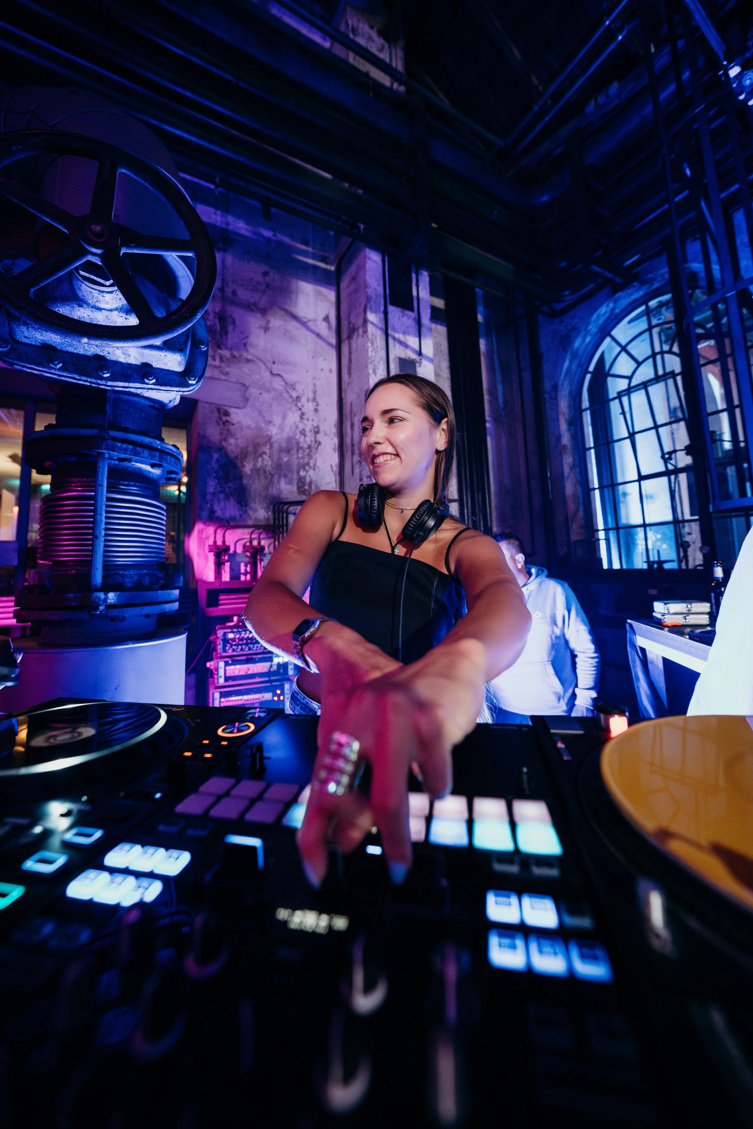 Female DJ wearing headphones, smiling, and operating a DJ controller in a dimly lit nightclub with purple and blue lighting.