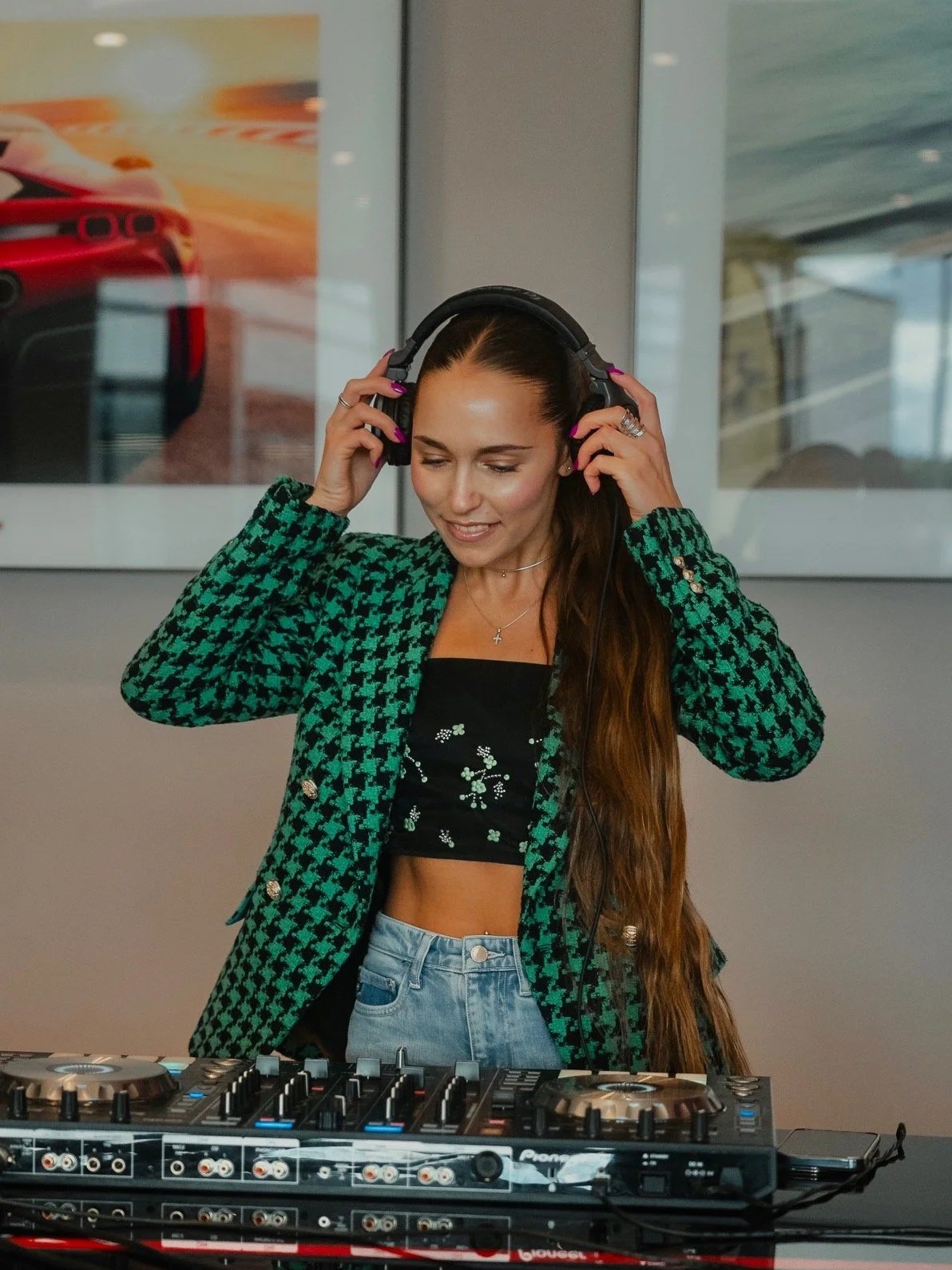 A woman with long brown hair, wearing a green and black houndstooth blazer over a black crop top with floral embroidery, is adjusting her large black headphones while standing behind DJ equipment.
