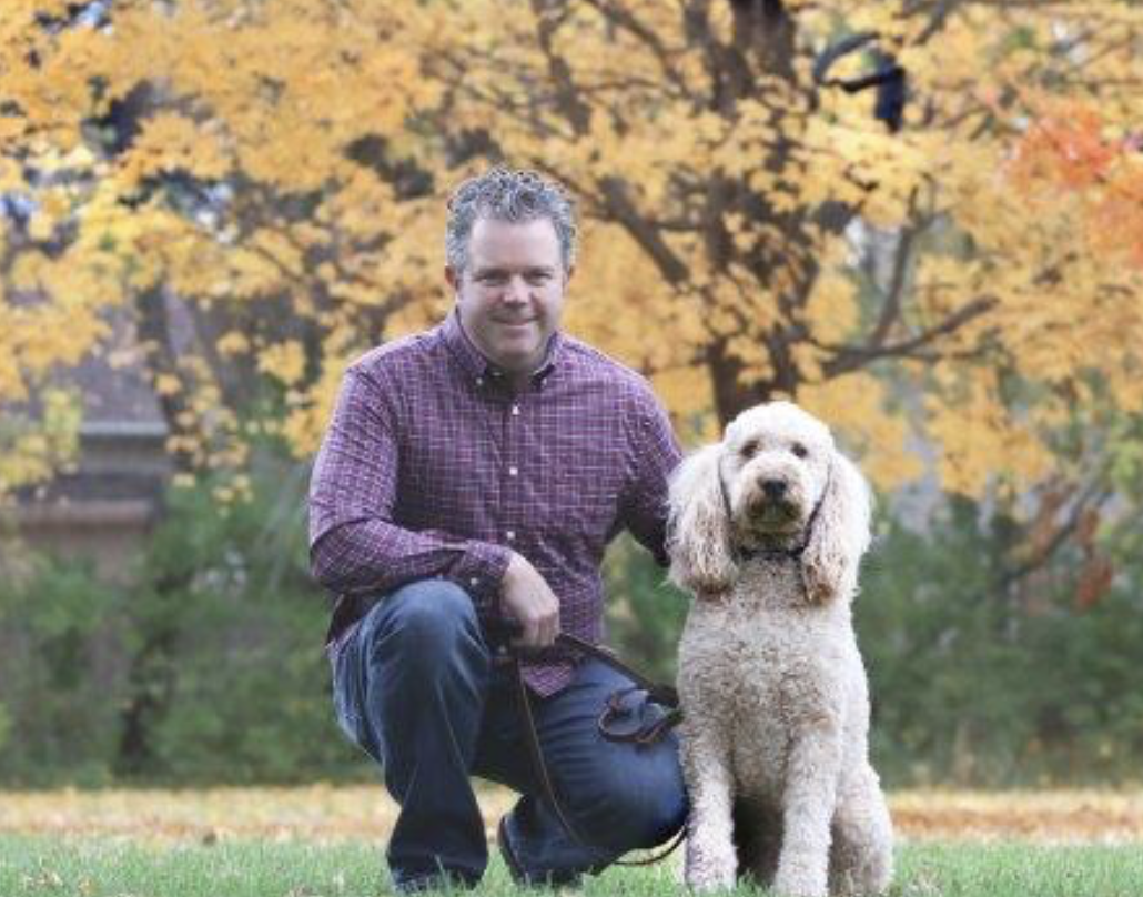 A man with curly hair, wearing a purple checkered shirt, kneeling beside a light-colored, curly-haired dog in a park with autumn trees and orange leaves in the background.