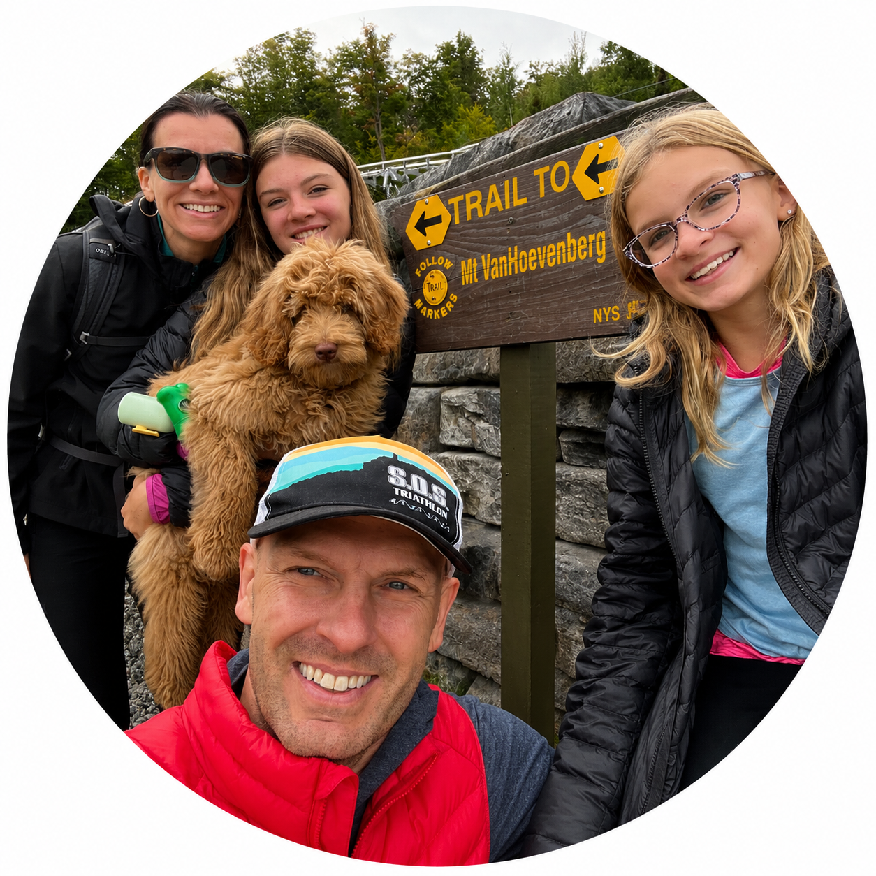 A family of four with a dog taking a selfie during a hike near a trail sign in a forested area.