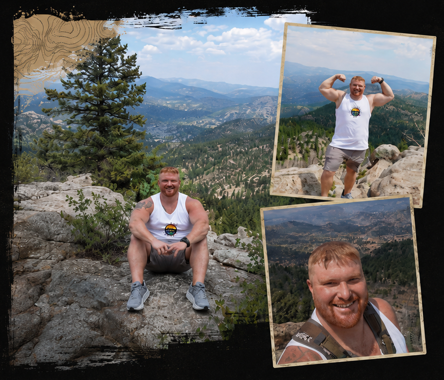 A man with red hair and a beard, wearing a white tank top and gray shorts, is hiking on a rocky mountain trail with a scenic valley in the background. He appears to be smiling and taking in the views.