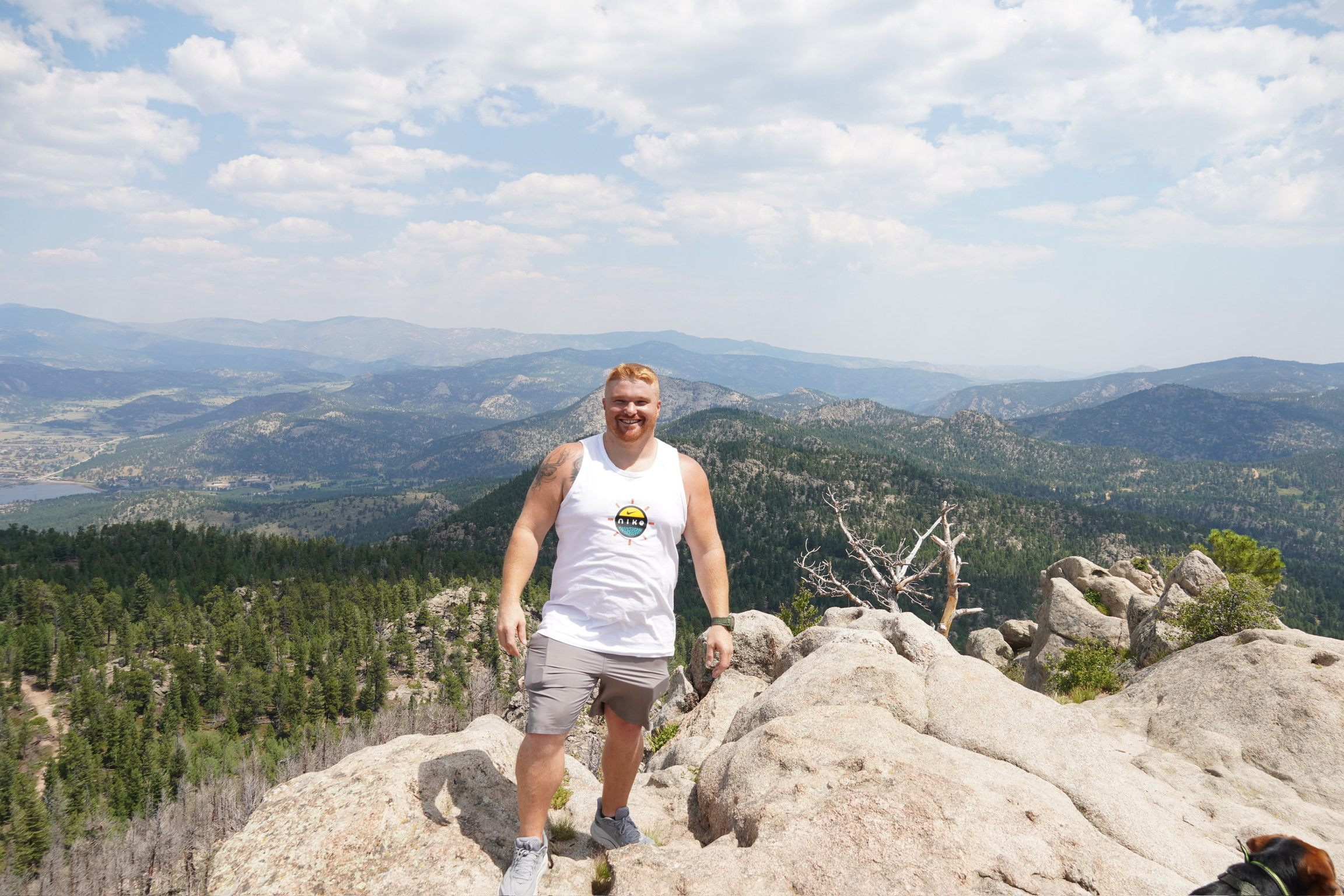 A man with red hair and a beard, wearing a white sleeveless shirt and gray shorts, standing on rocks with a mountainous landscape and blue sky with clouds in the background.