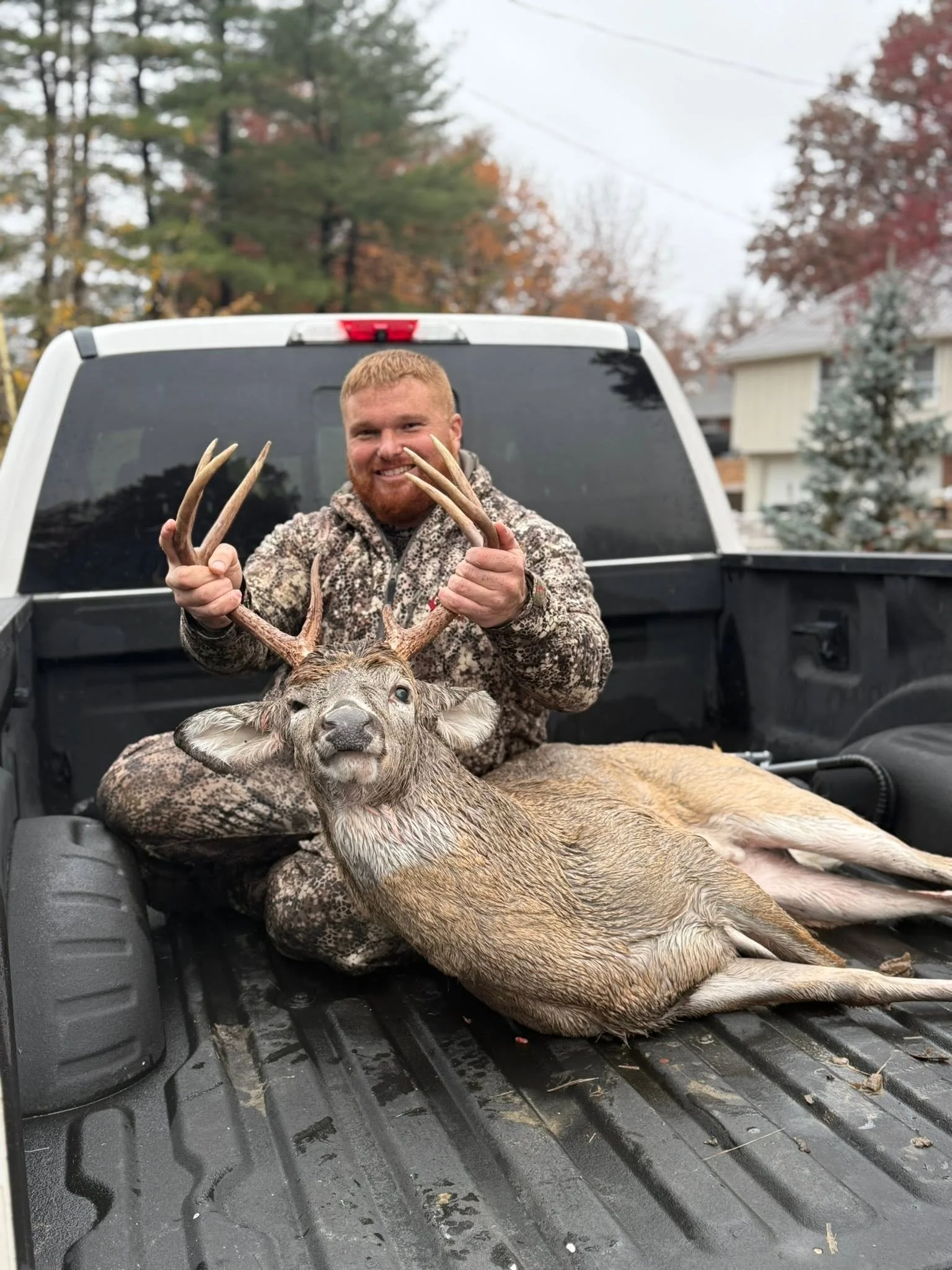 A smiling man with red hair and beard, wearing camouflage clothing, sitting in the back of a pickup truck, holding up the antlers of a large deer with a white tail, during daytime with trees and houses in the background.