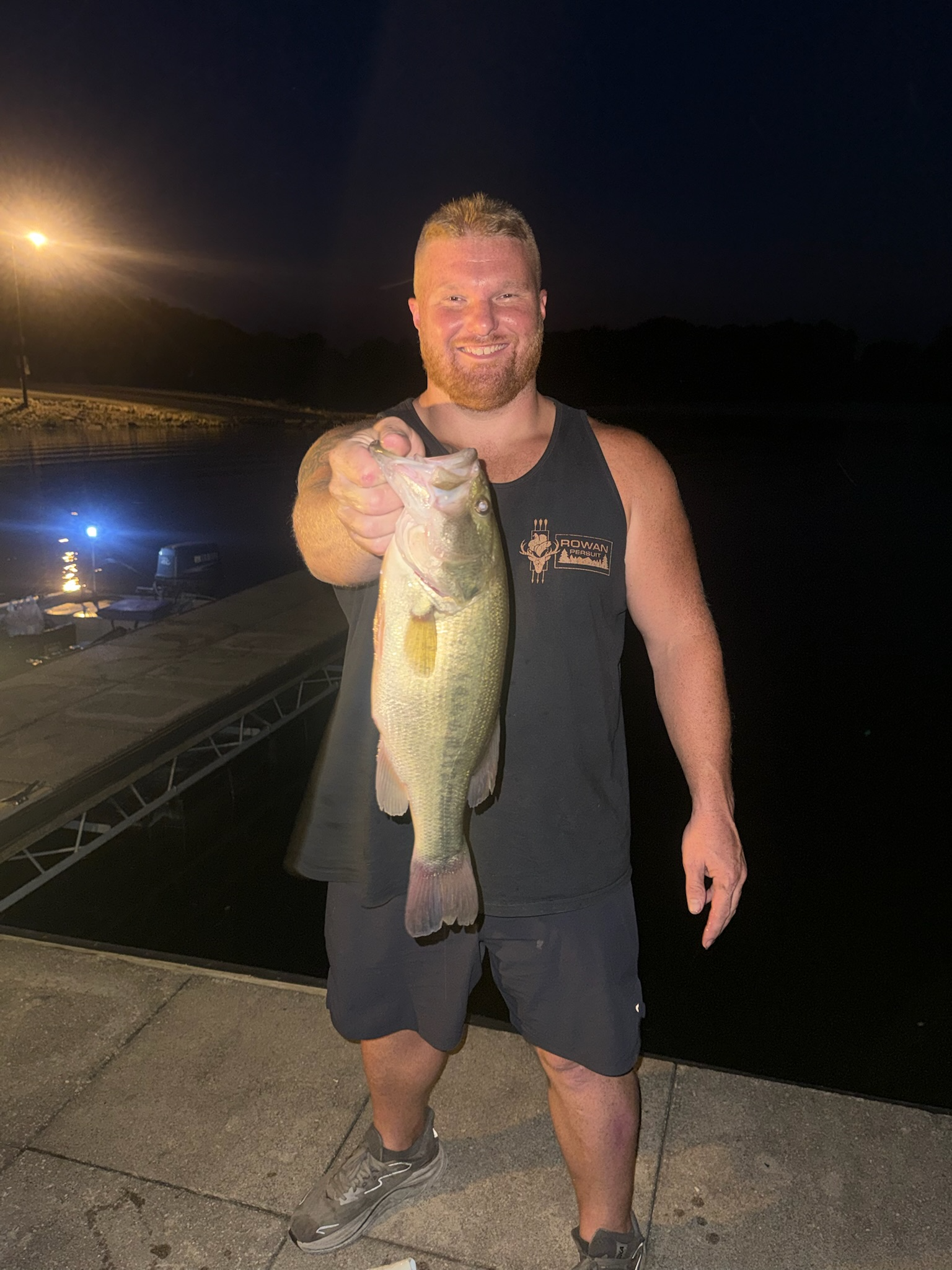 A man with a beard and short hair smiling while holding a large fish in nighttime setting near a dock or marina, with a boat and water in the background.