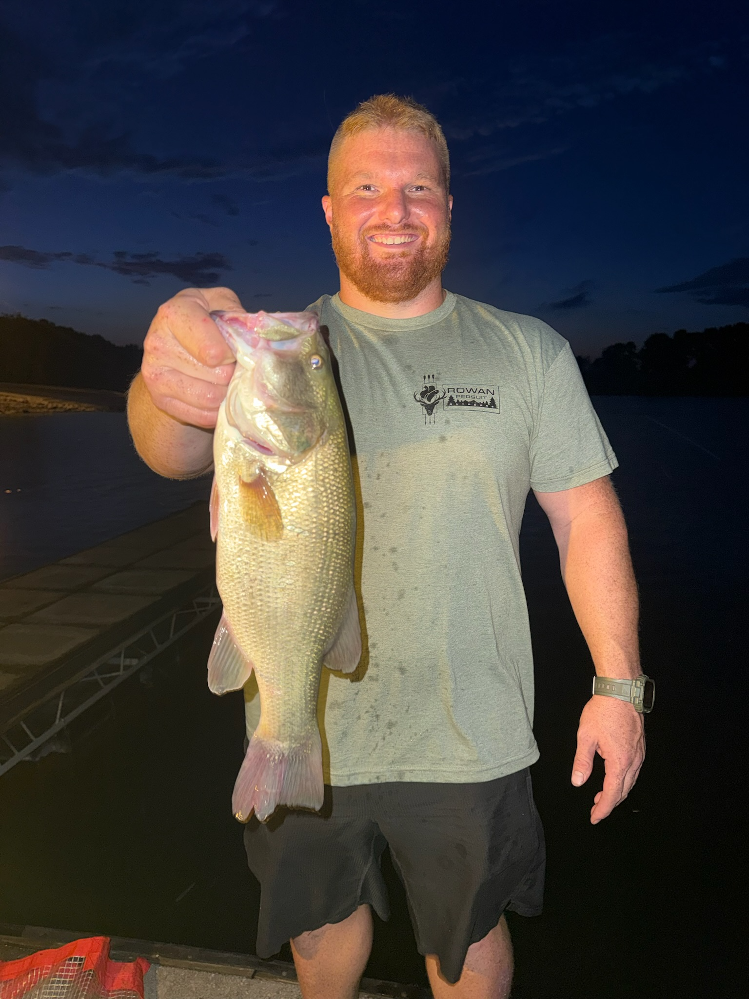 A man with red hair and a beard smiling while holding a largemouth bass fish during dusk or early night, with a dark sky background.