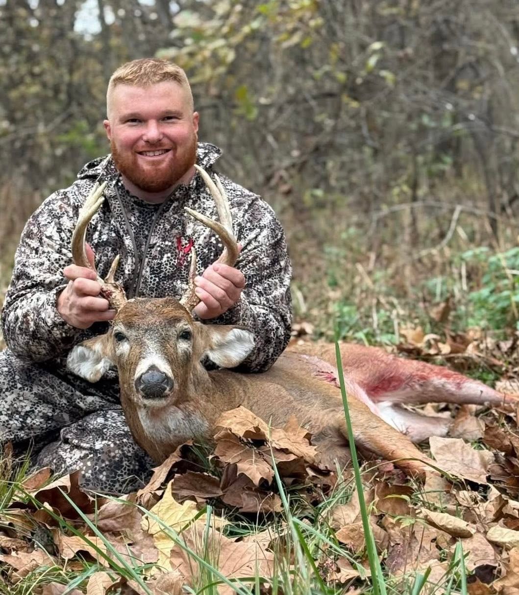 A man in camouflage clothing kneels in the woods, holding the antlers of a slain deer lying on a bed of fallen leaves.