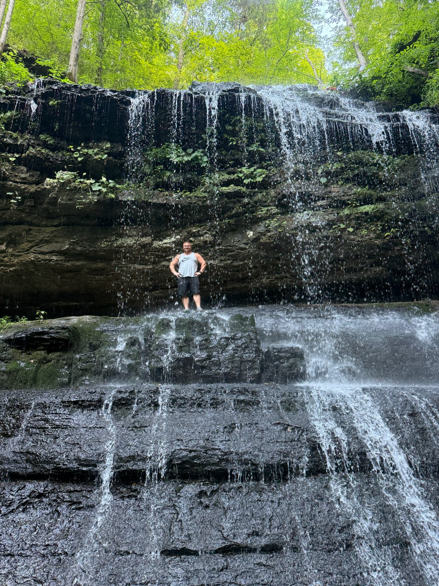 A person standing under a waterfall in a lush green forest with sunlight filtering through the leaves.