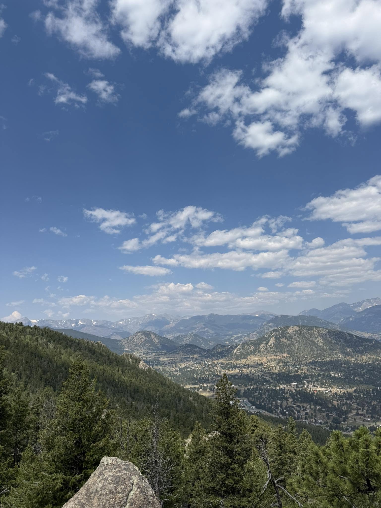 View of a mountainous landscape with a forest in the foreground and a valley below, under a blue sky with scattered white clouds.