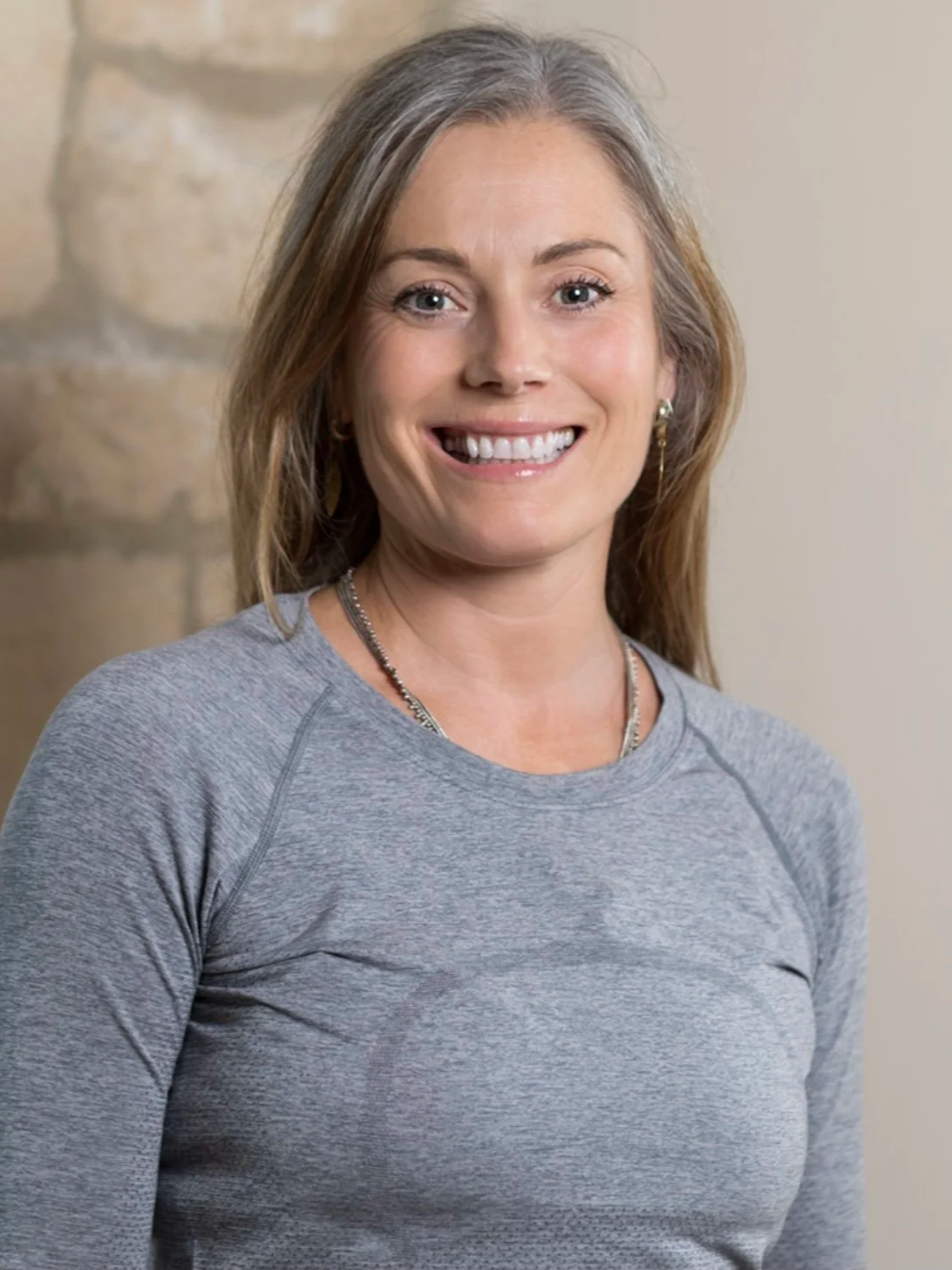 A woman with long, light brown hair, smiling, wearing a gray athletic shirt and jewelry, standing indoors in front of a stone wall and neutral-colored wall.