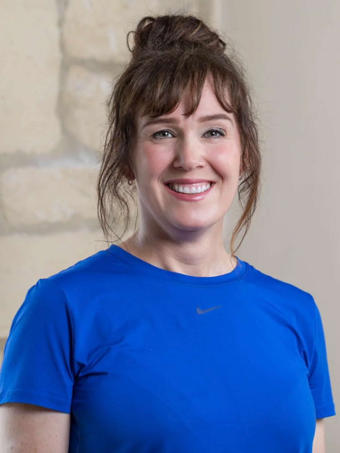 A smiling woman with red hair styled in a messy bun, wearing a blue Nike athletic shirt, standing indoors against a beige wall with stone accents.