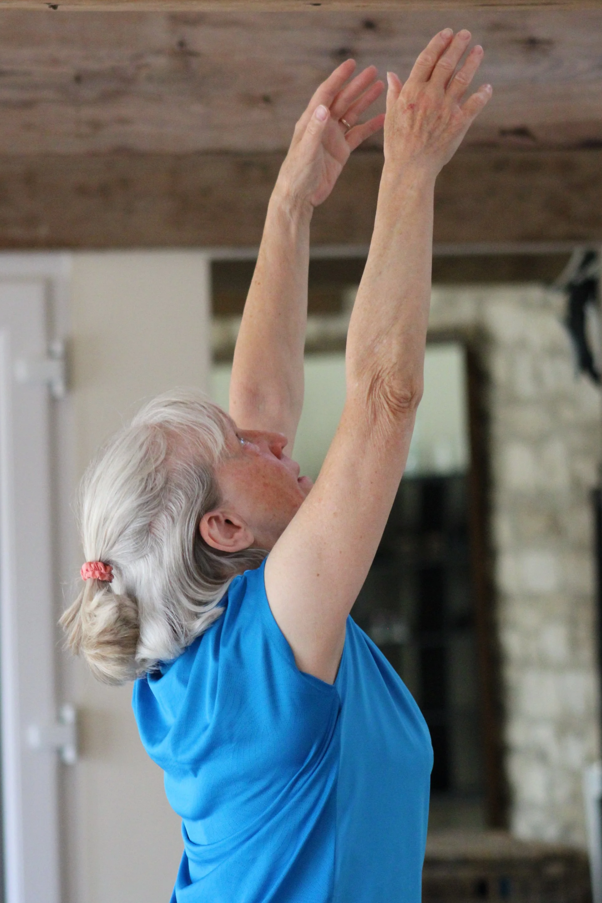 Old woman with gray hair tied back practicing Pilates movement, wearing a blue shirt.