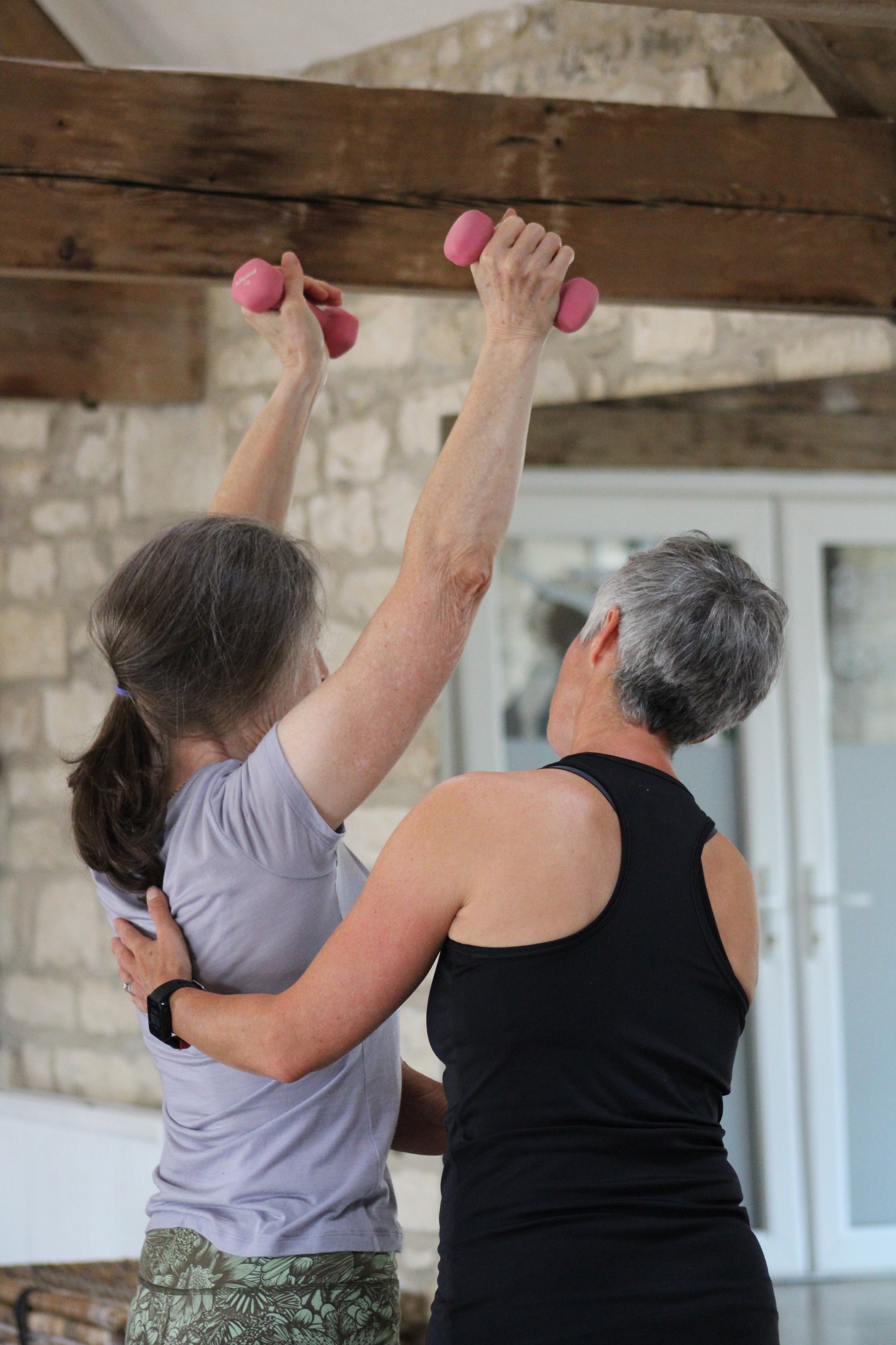 Two women in shot, one woman exercising with pink dumbbells wearing a purple t-shirt and patterned shorts and the other wearing ablack tank top, in a room with a stone wall and wooden beams.