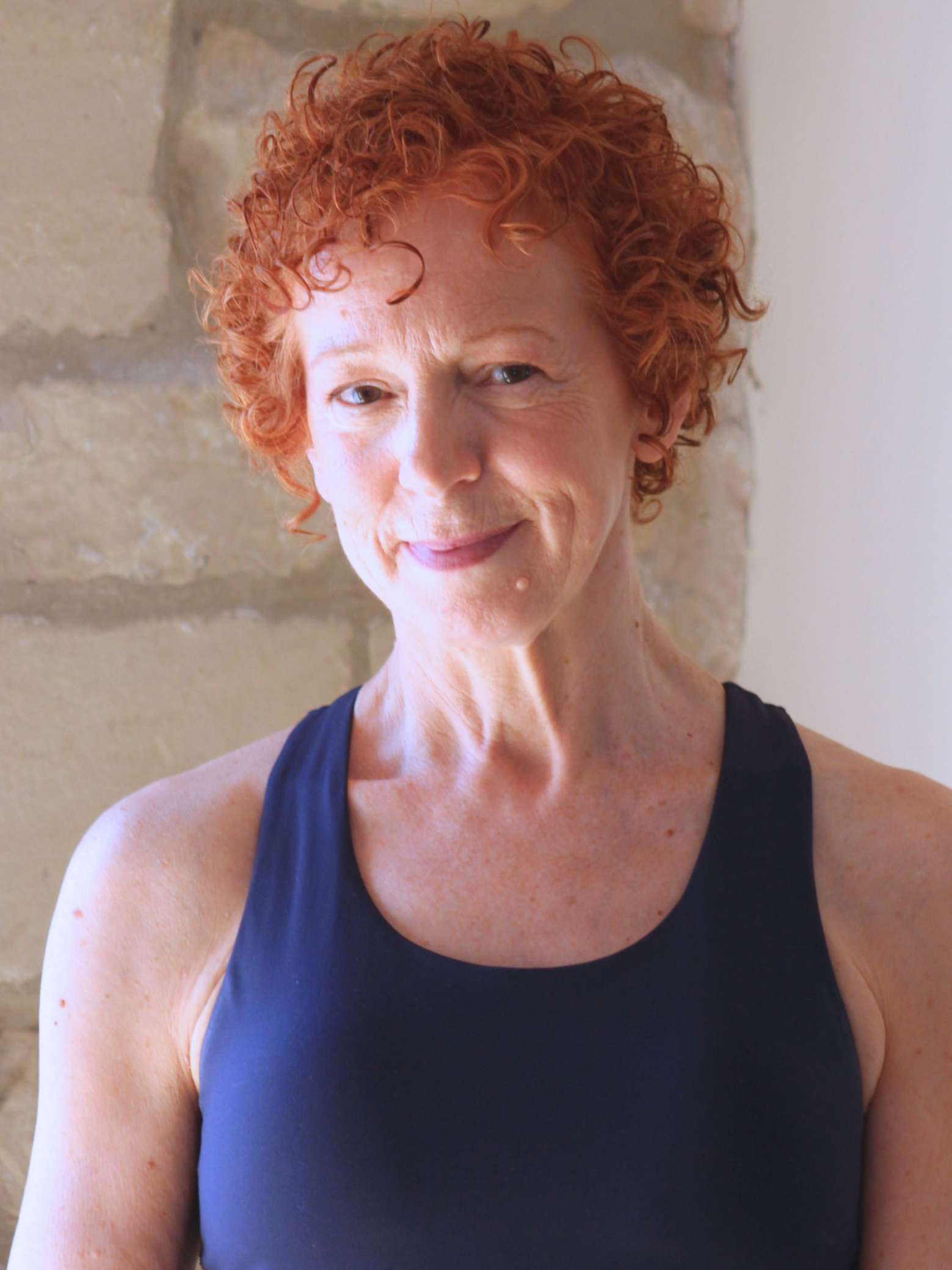 A woman with short, curly red hair and fair skin is wearing a black sleeveless top. She is standing against a light-colored brick wall. Her expression is gentle and she appears to be smiling slightly.