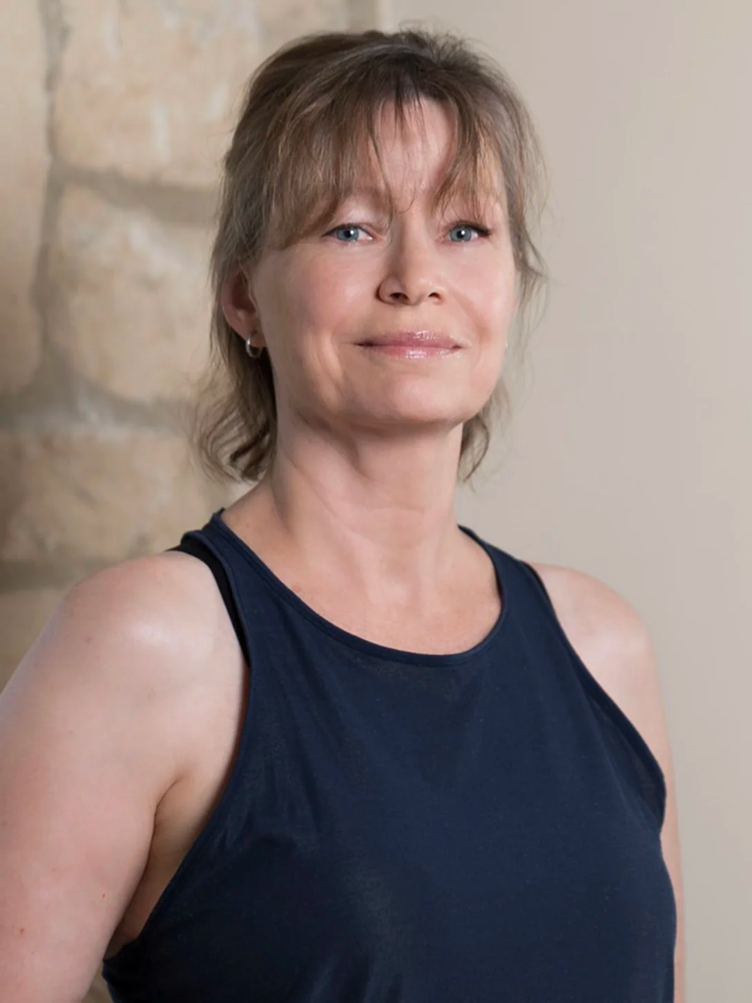 A woman with short, light brown hair, blue eyes, and wearing a dark sleeveless top, standing indoors near a stone wall.
