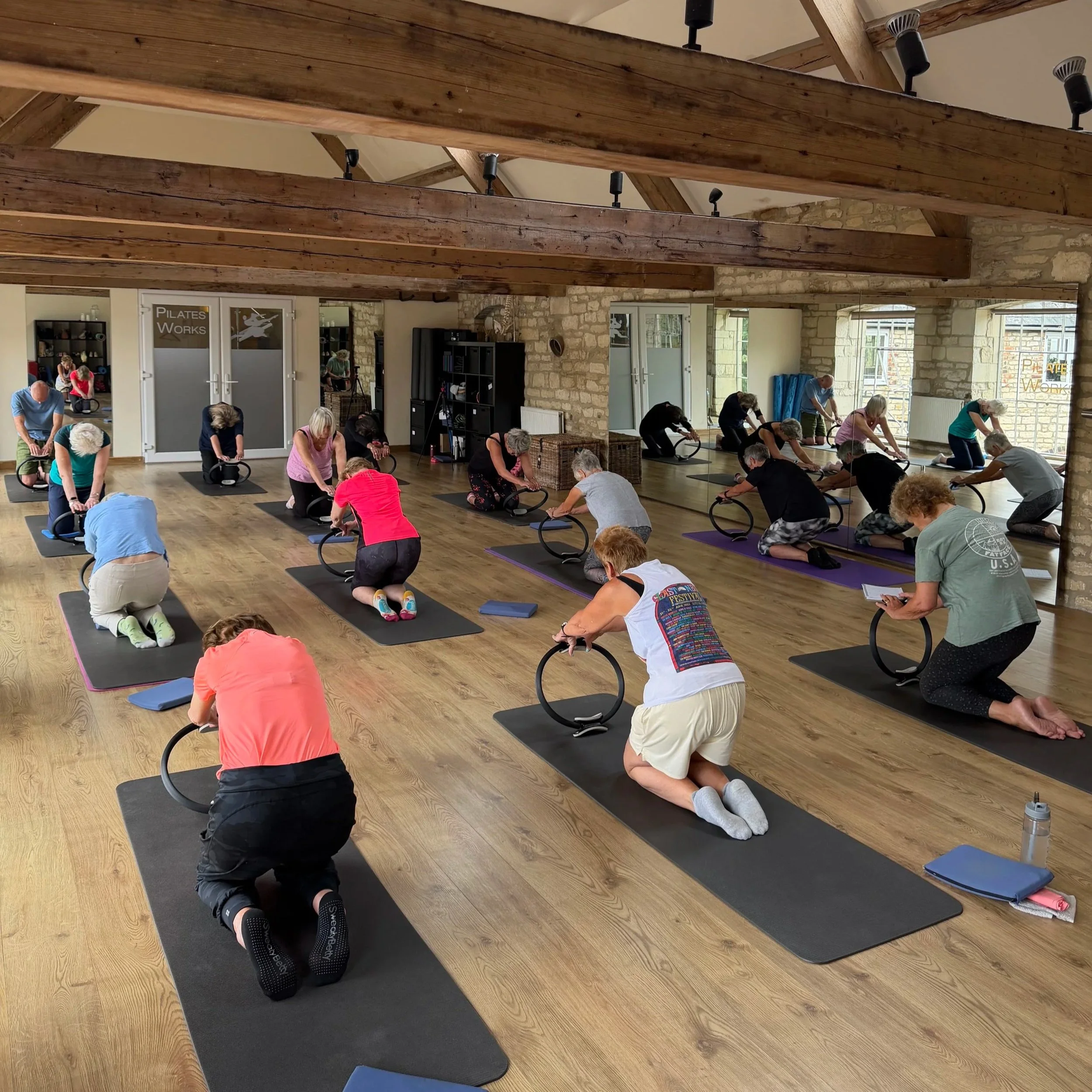 A group of people participating in a Pilates class in a spacious studio with wooden beams and brick walls. They are kneeling on mats, using small exercise rings, and wearing comfortable workout clothing.