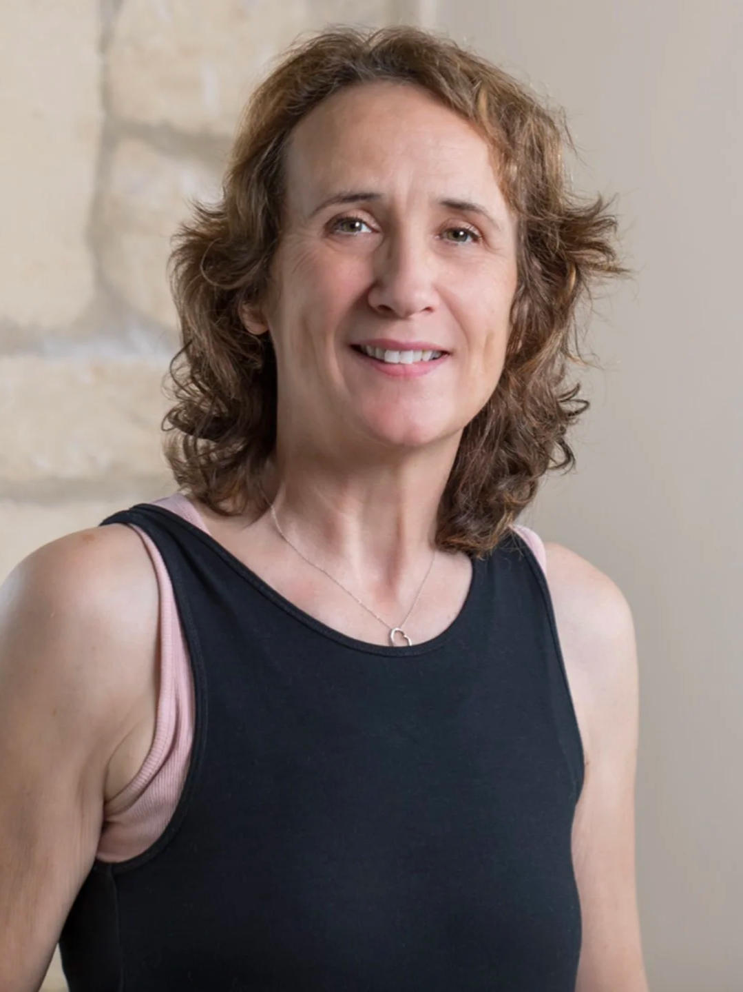 A woman with curly brown hair wearing a black sleeveless top and a silver necklace, smiling at the camera, standing indoors against a light-colored wall with a stone feature.