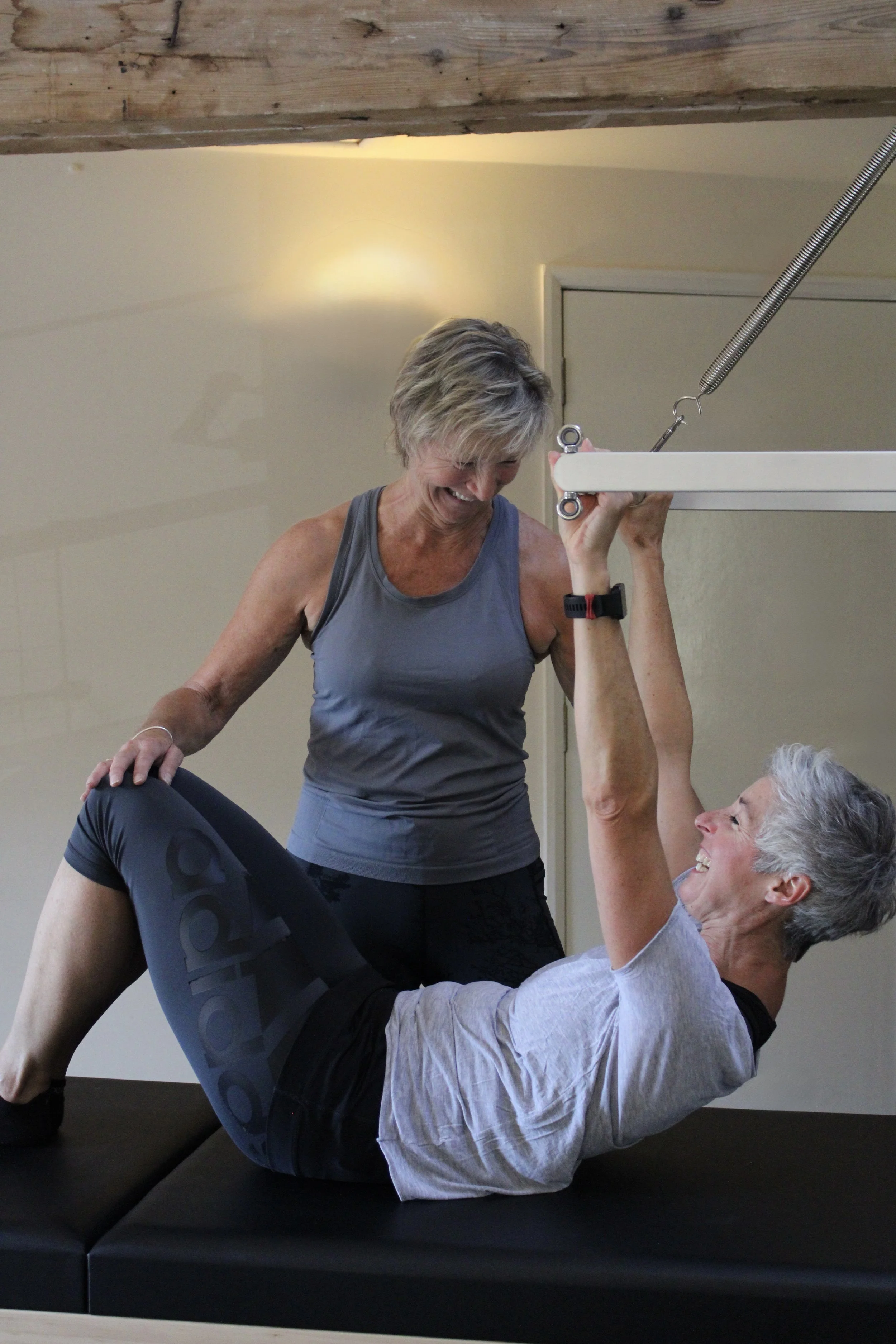 Two women using Pilates equipment, with one woman lying on a padded surface while the other woman checks her form and provides guidance at a 1-1 level.