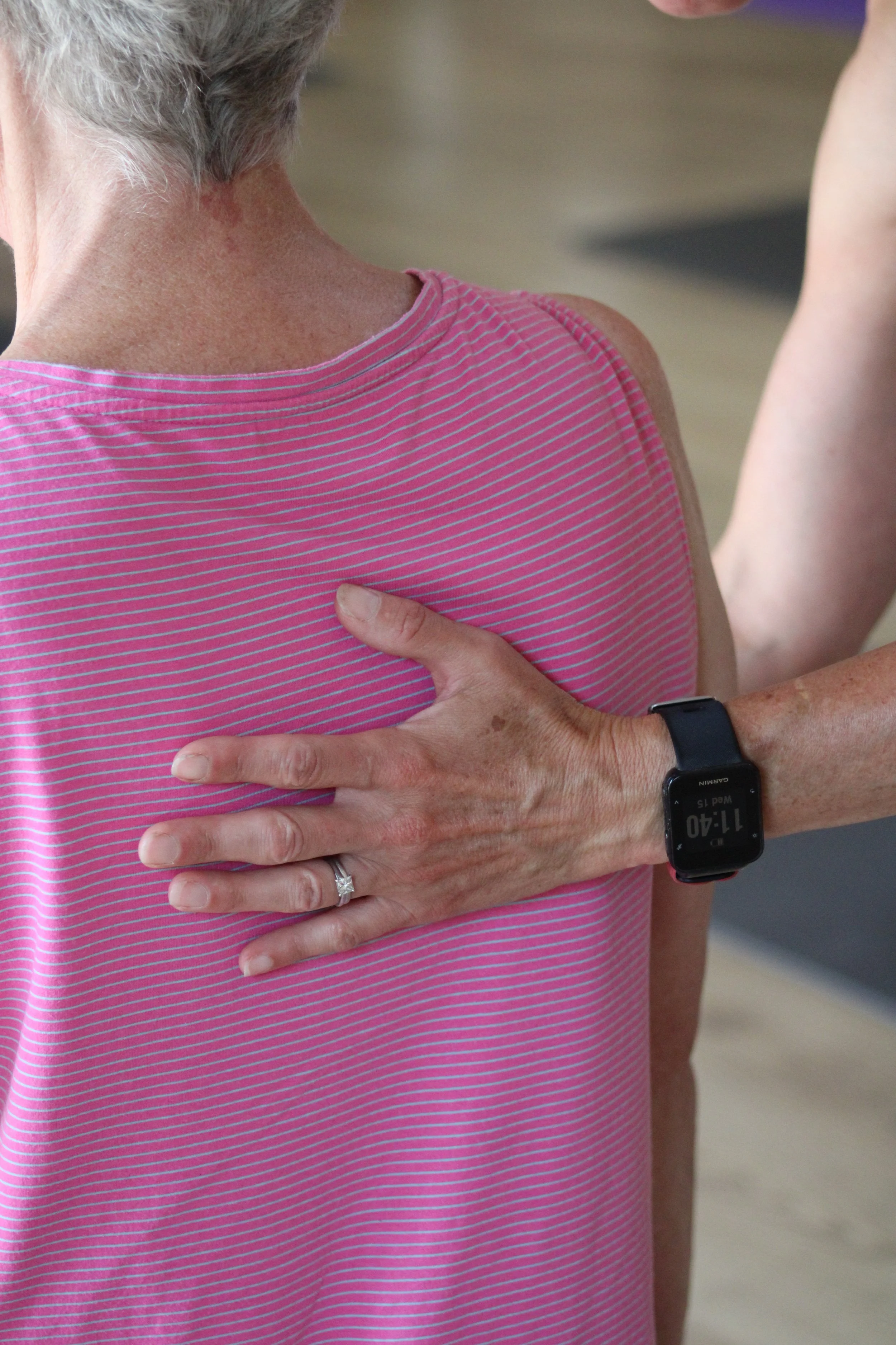 A person with short gray hair wearing a pink and gym vest, our instructor is providing guidance on best form using her hand on the client's back. She is wearing a black wristband.