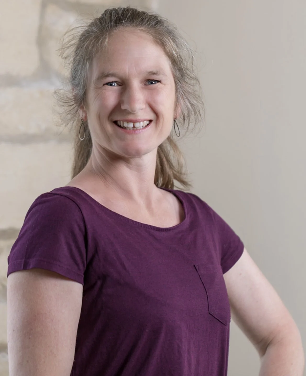 Smiling woman with light brown hair wearing a purple shirt, standing against a beige wall.