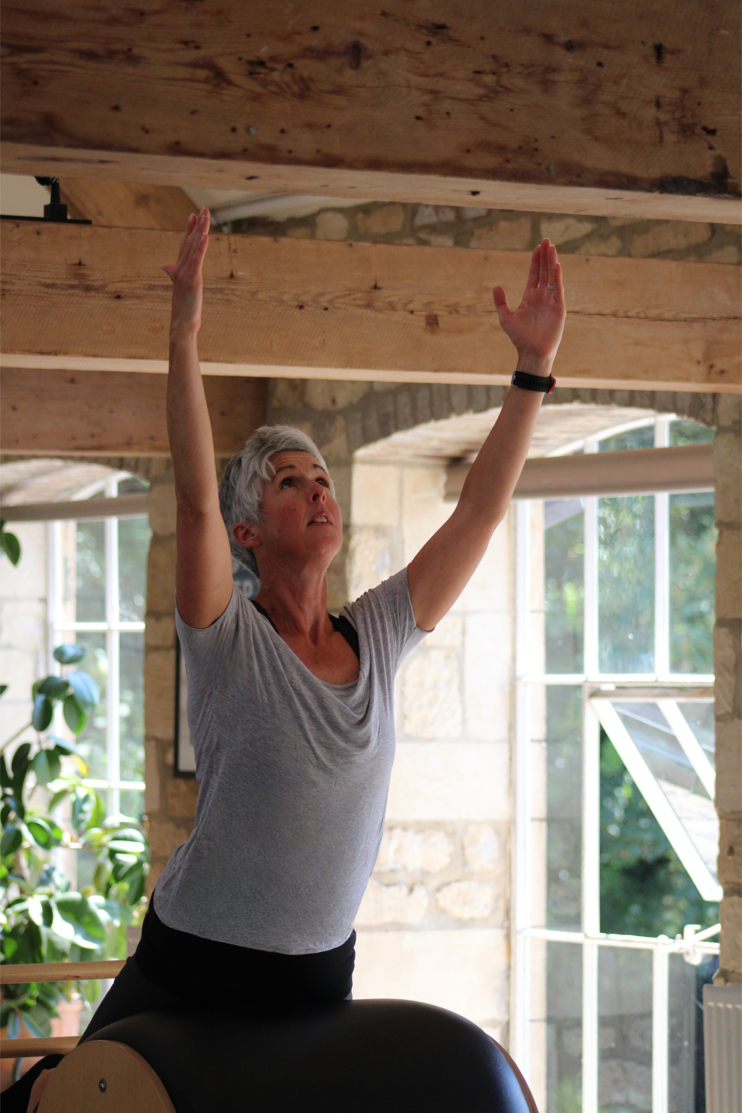 Woman with short gray hair practicing Pilates on a machine in a bright room with large windows and exposed brick walls.