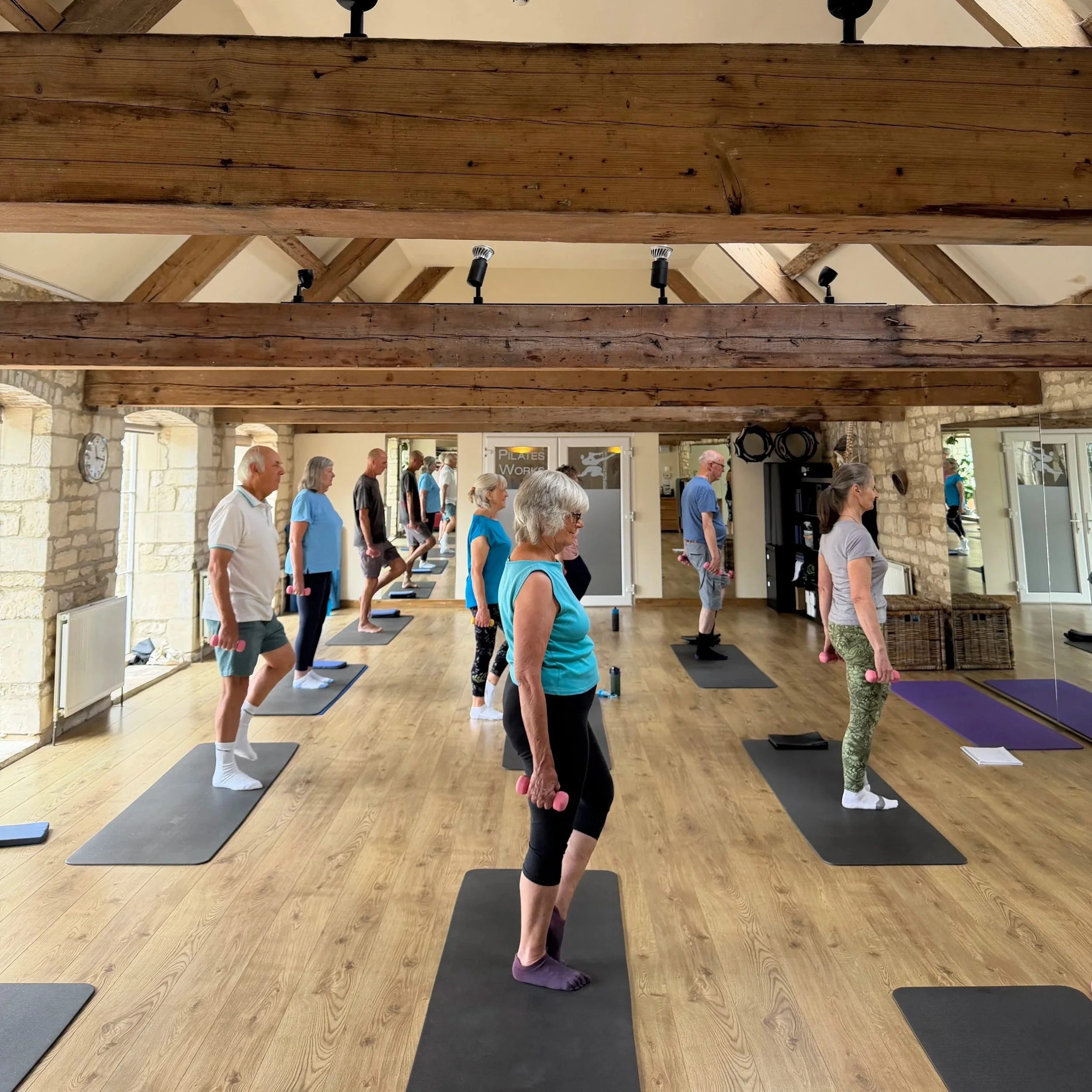 Group of people participating in a Pilates class in a room with wooden beams and stone walls, some holding small pink weights, standing on Pilates mats.