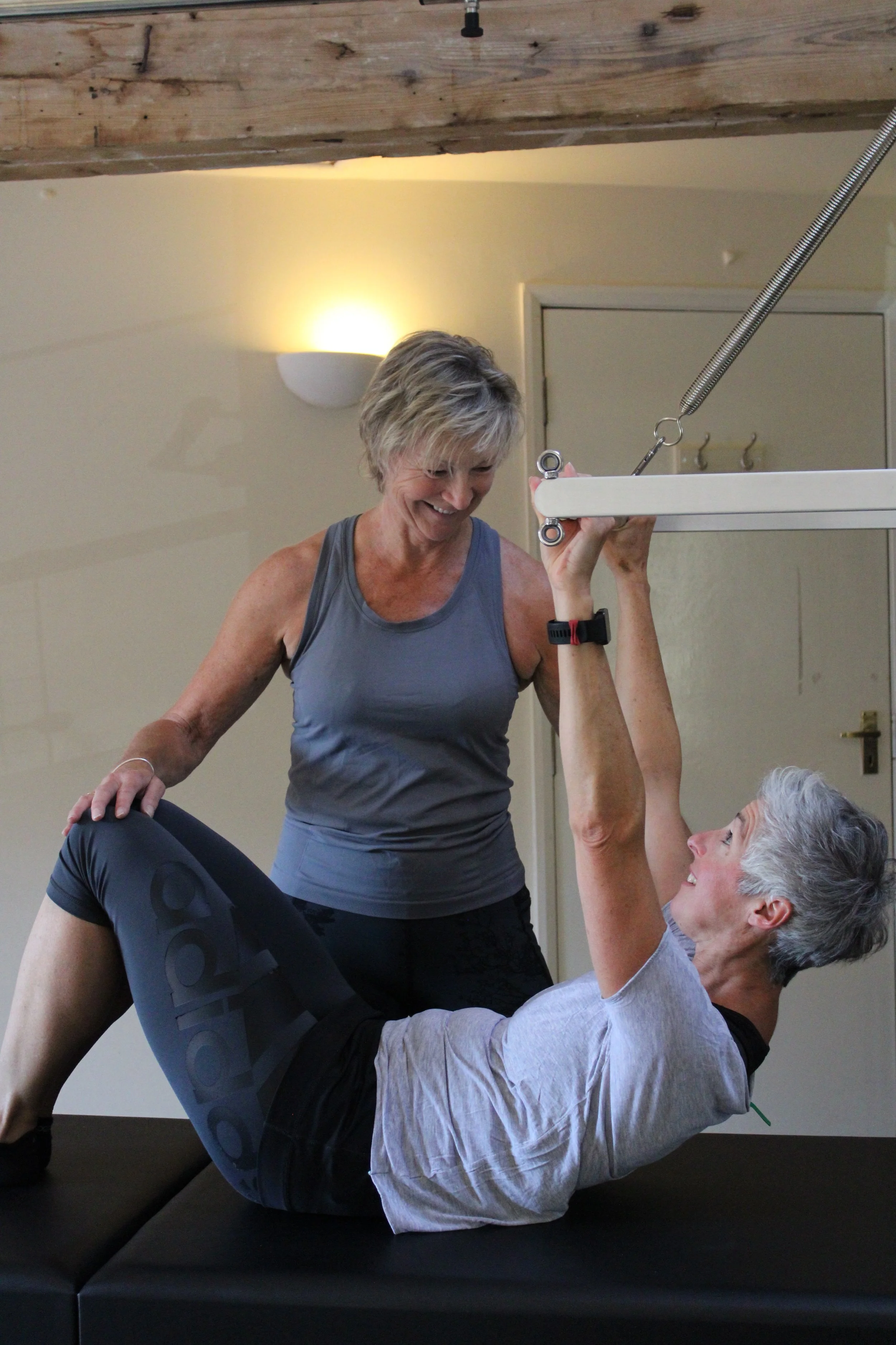 Two women using Pilates equipment, with one woman lying on a padded surface while the other woman checks her form and provides guidance at a 1-1 level.