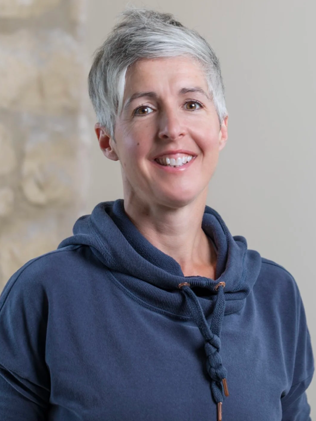 A smiling woman with short gray hair wearing a navy blue hoodie, standing indoors with a neutral background and part of a stone wall visible.