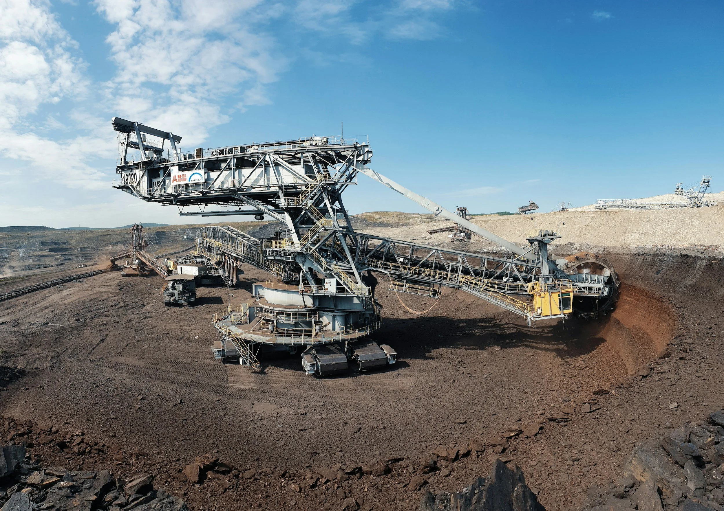 Open-pit mining site with large conveyor system and heavy machinery moving earth, under a blue sky with some clouds.