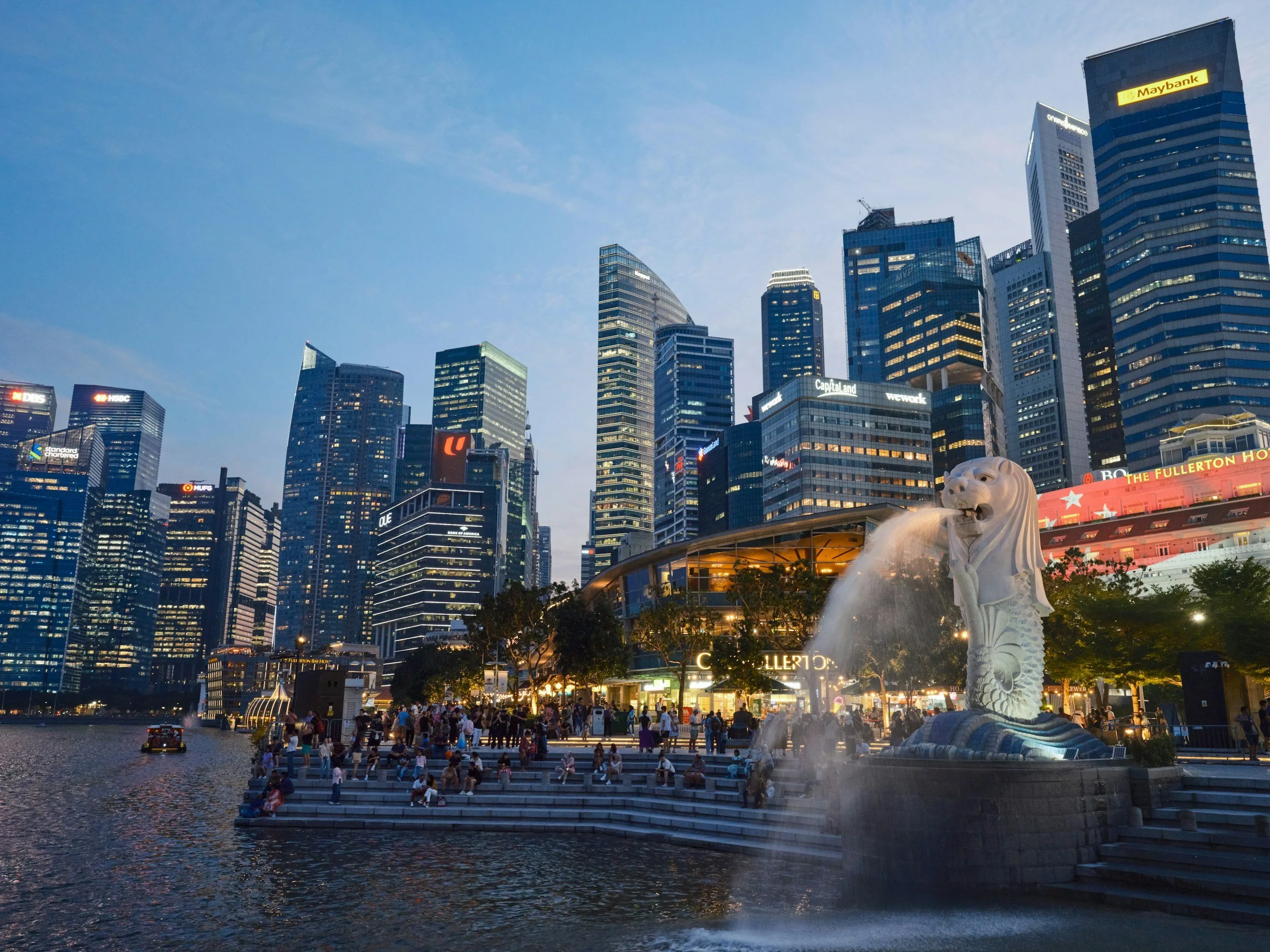 Nighttime cityscape of Singapore with tall skyscrapers, the Merlion statue in the foreground, and people gathered at the waterfront.
